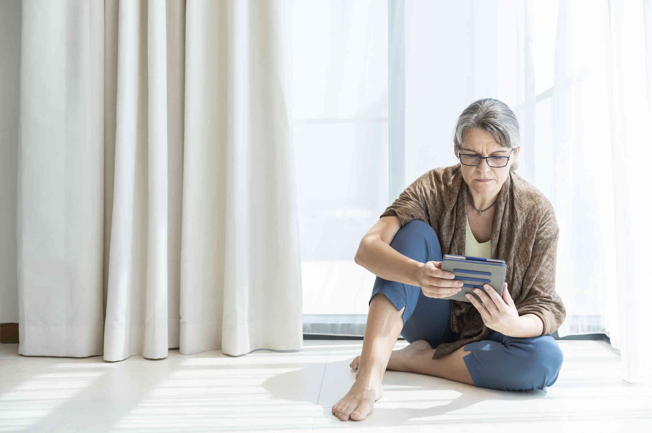 A middle aged woman with glasses sits on the floor in her home and looks at the tablet in her hands.