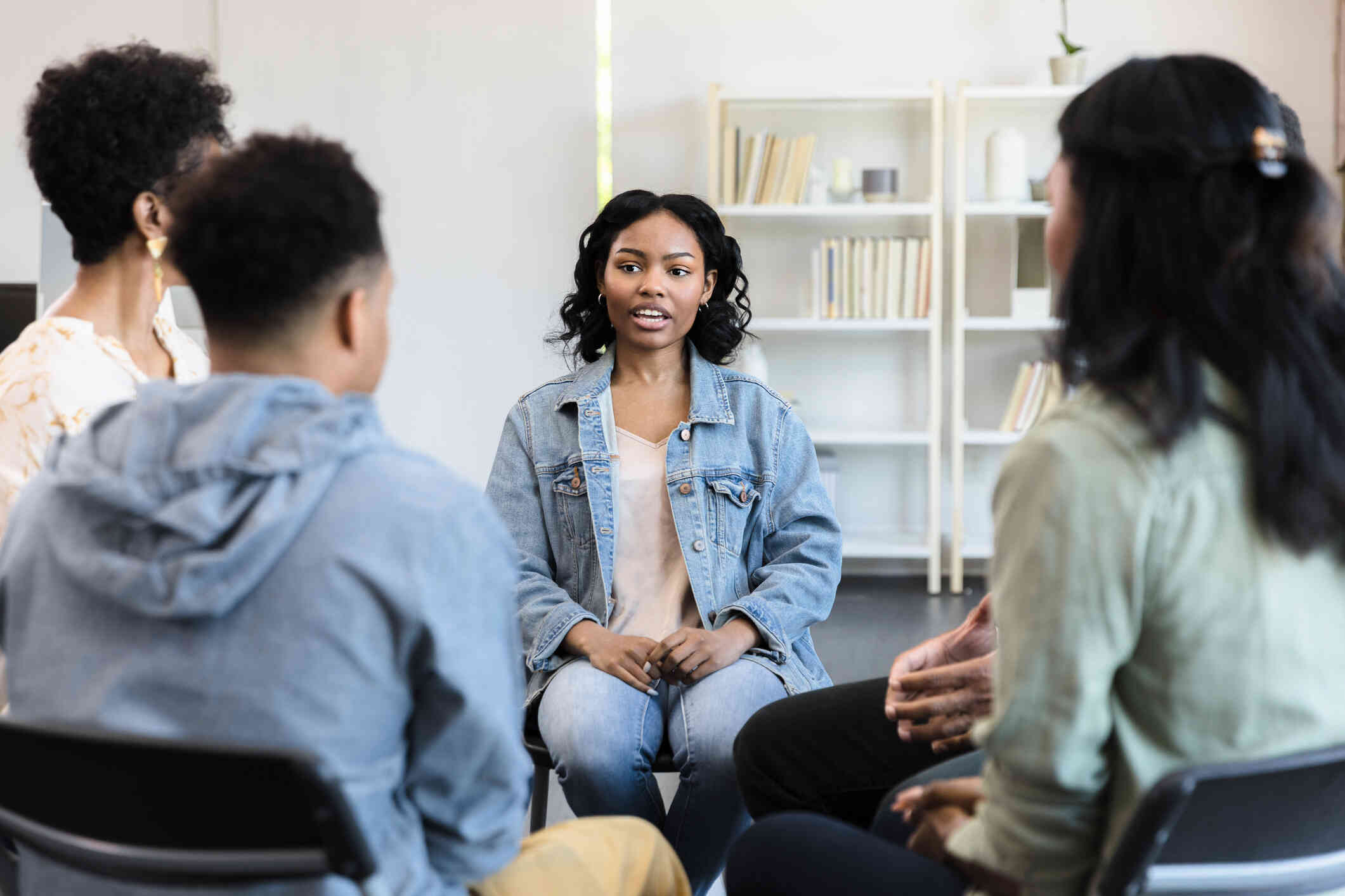 A group of adults sit in chairs in a therapy circle as a woman talks.