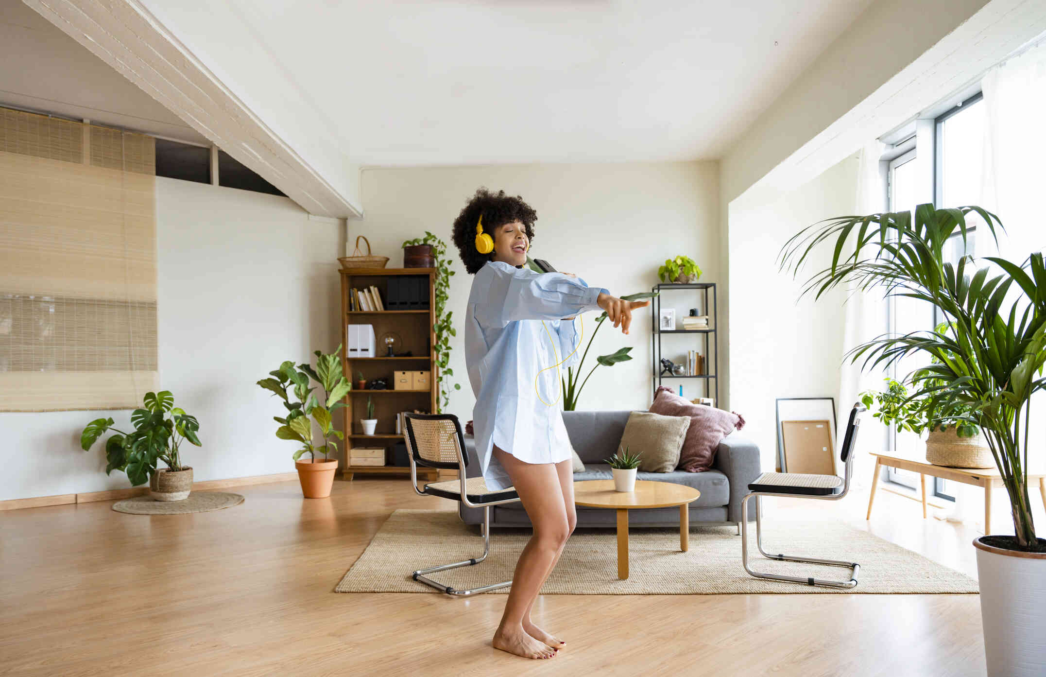 A woman in an oversized button down shirt dances happily by herself in her livingroom while wearing yellow headphones.