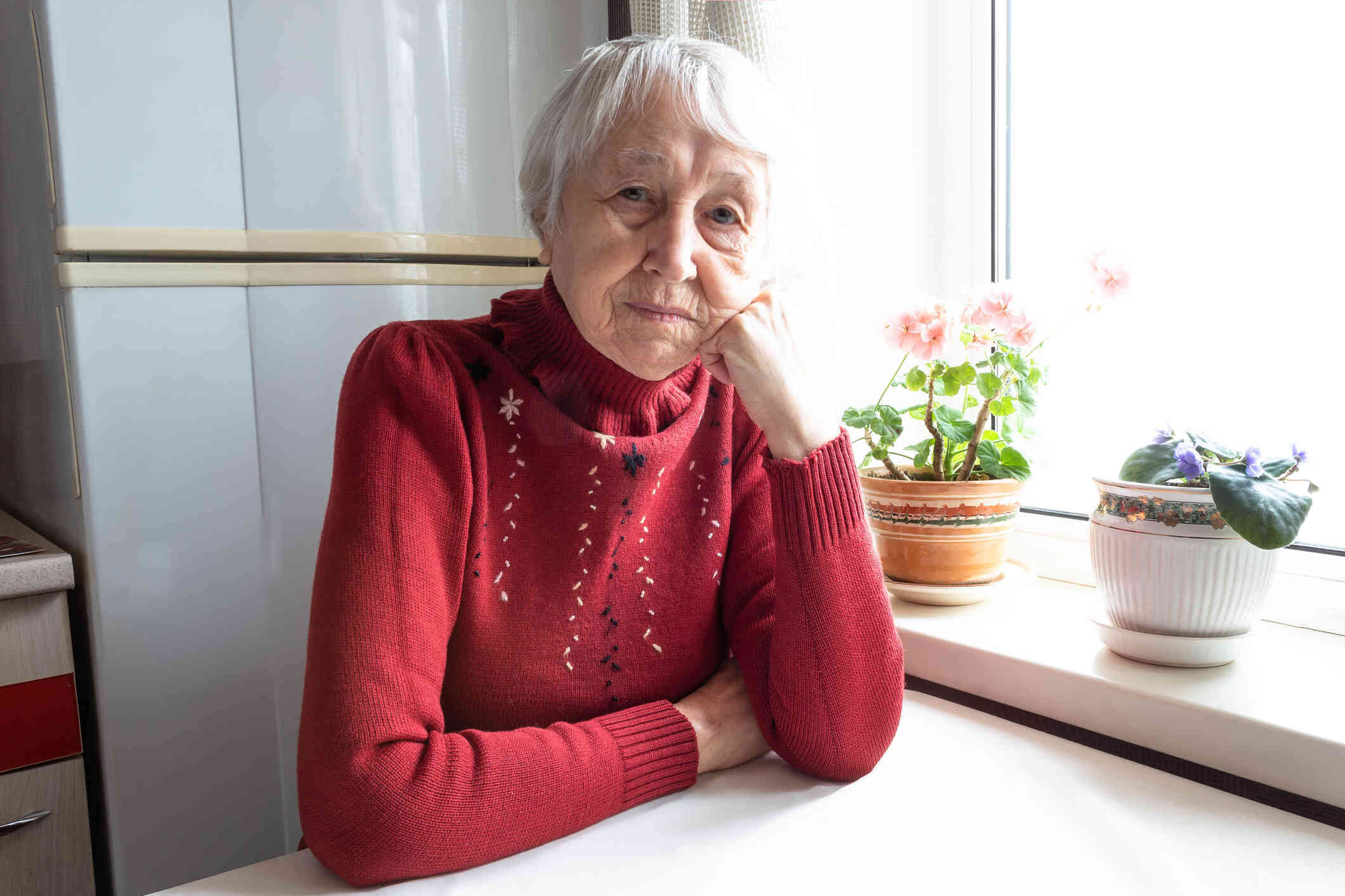 An elderly woman in a red sweater sits at her kitchen table and rests her head on her closed hand while looking at the camera with a sad expression.