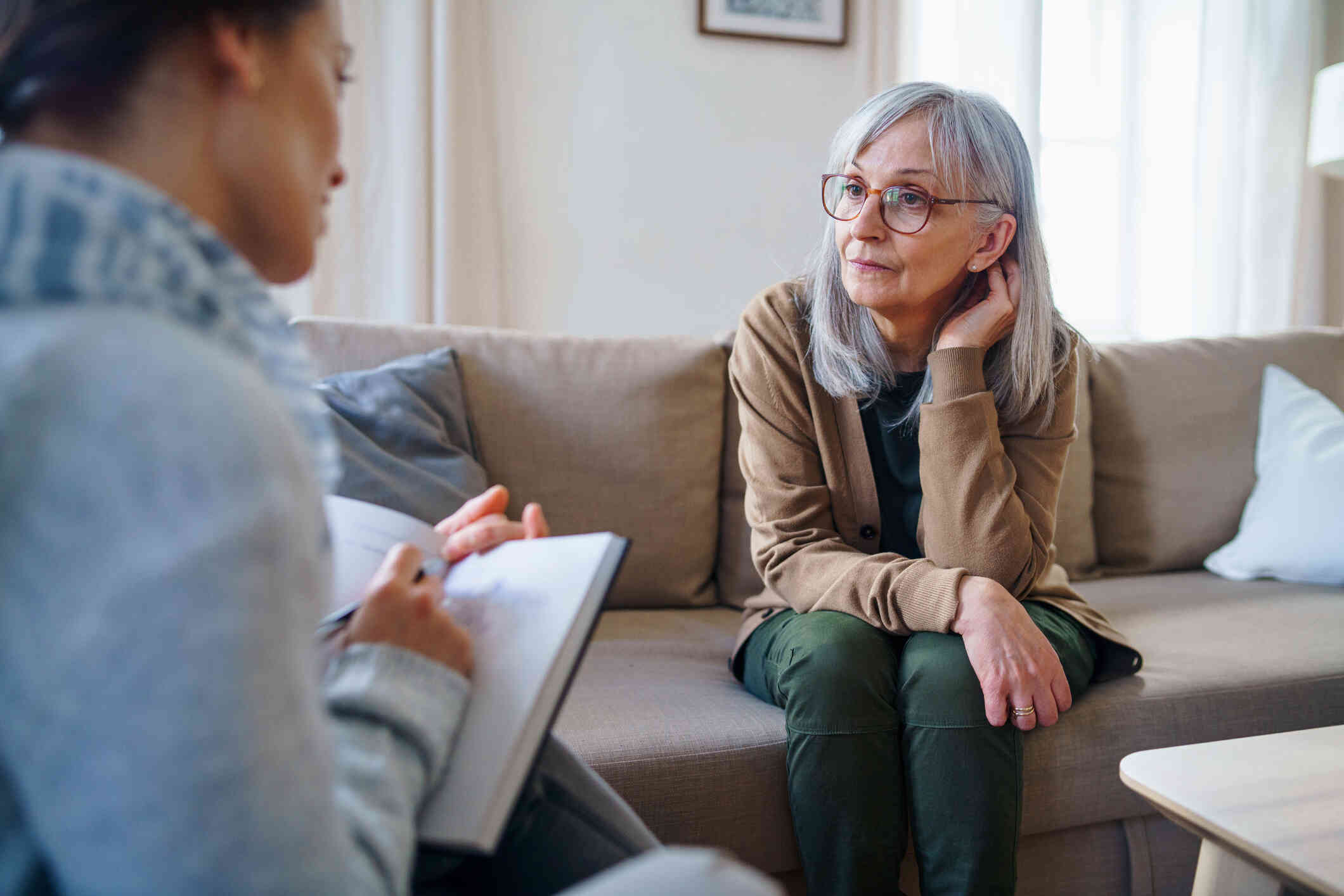 A mature woman with glasses sits hunched voer on the couch as she looks at the therapist sitting across from her with a worried expression.