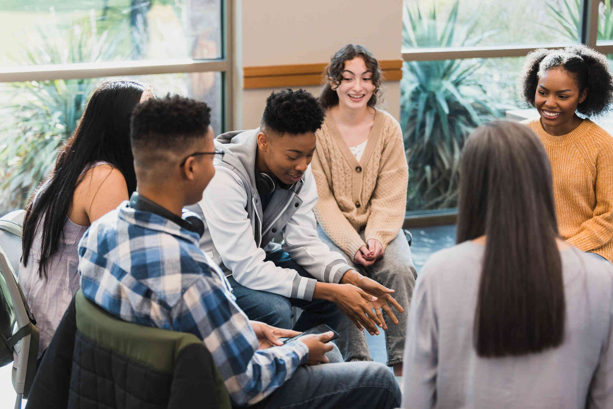 A group of young women and men with positive facial expressions sit around in a circle in chairs while having a conversation.