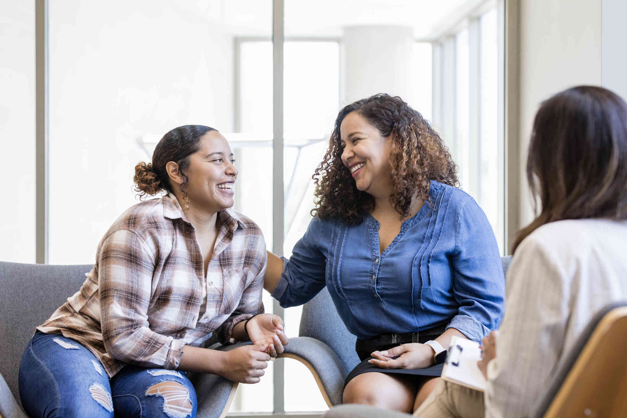 Two sibling sisters sit in chairs next to each other while laughing and smiling at one another.