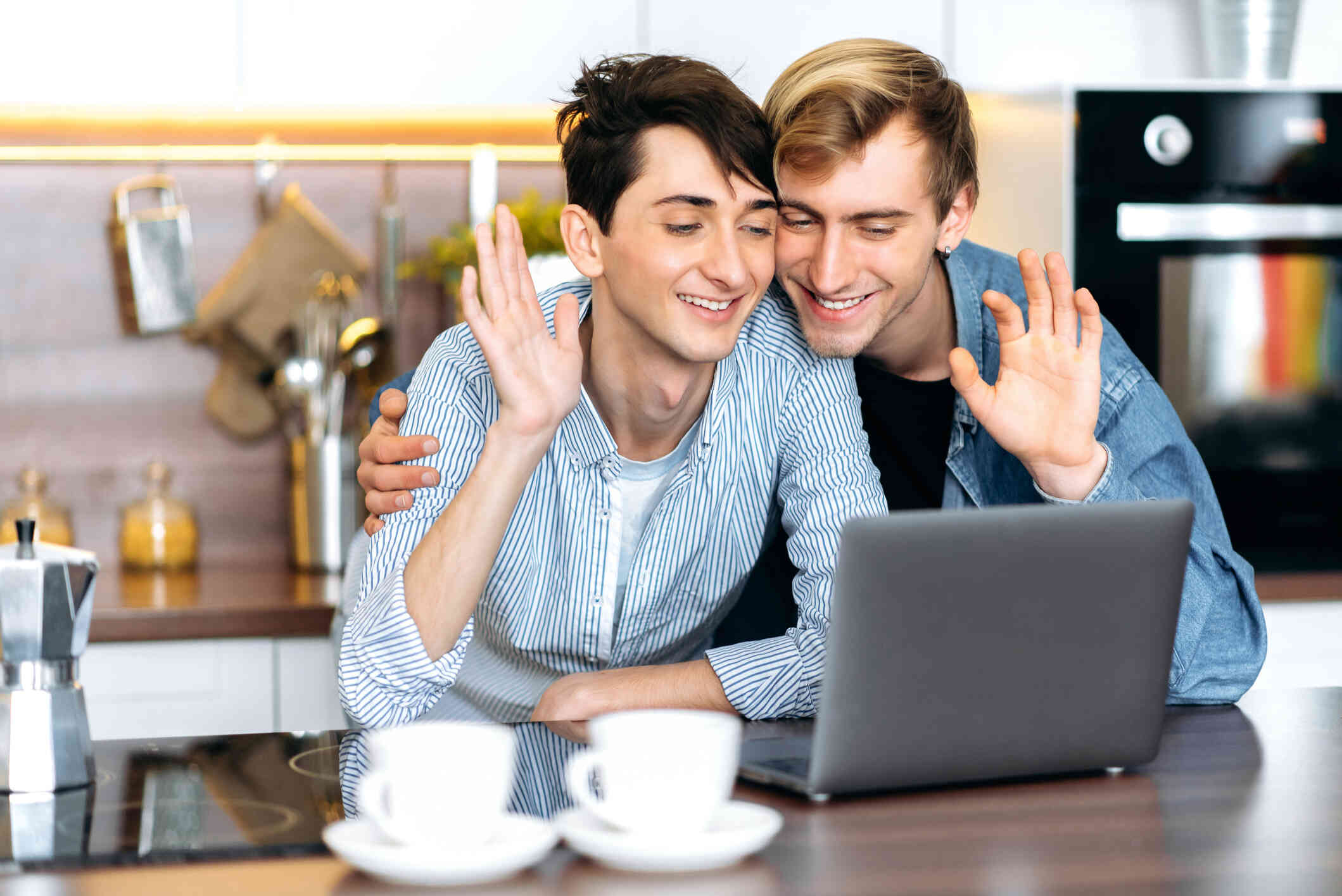 A couple seated at a table waves and smiles at the laptop screen.