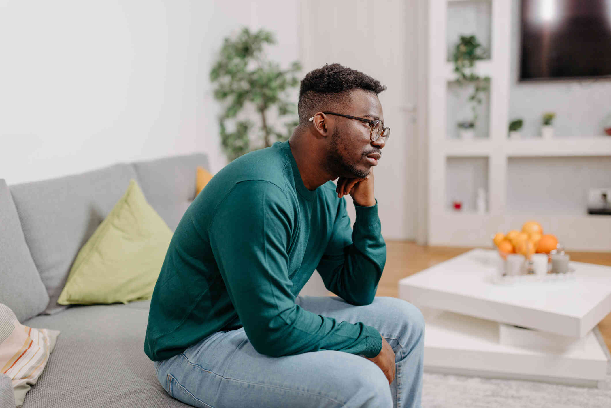 A man in a long sleeve green shirt sits hunched over on the couch and rests his head on his hand with a sad expression.