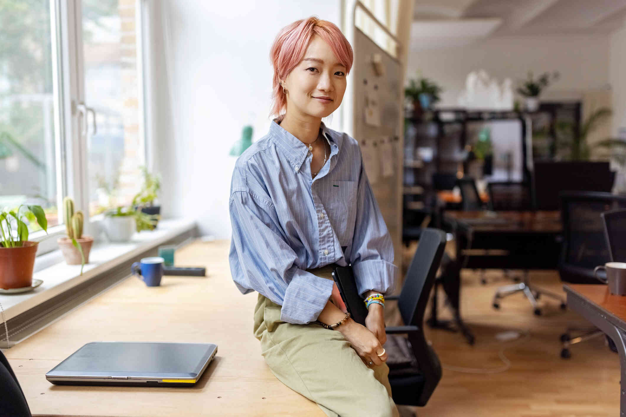 A woman with short pink hair leans against the counter in an office space and smiles softly at the camera while holding a closed laptop.