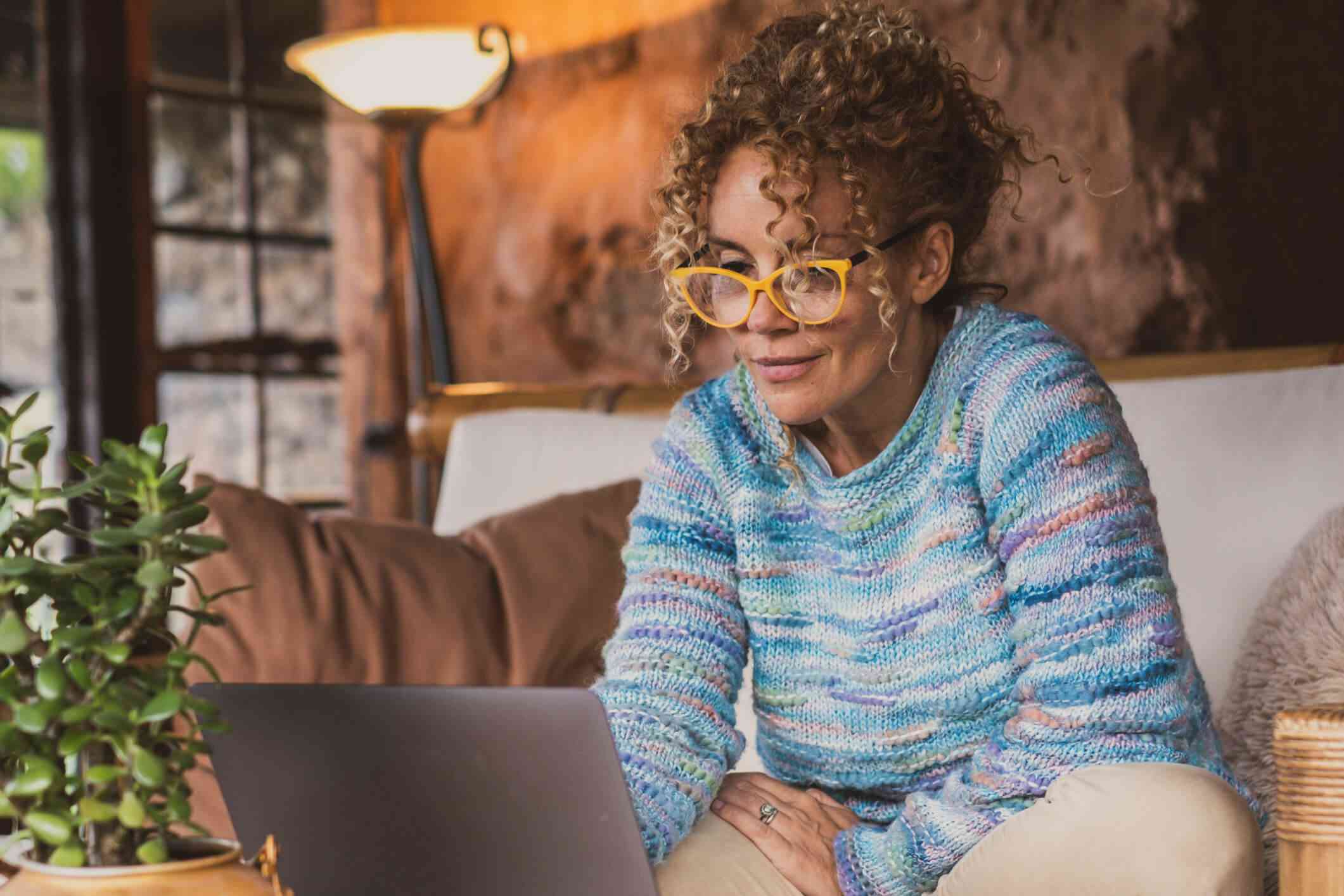 A woman wearing glasses and a colorful sweater sits hunched over on the couch while she looks at the laptop that is open on the coffee table infront of her.
