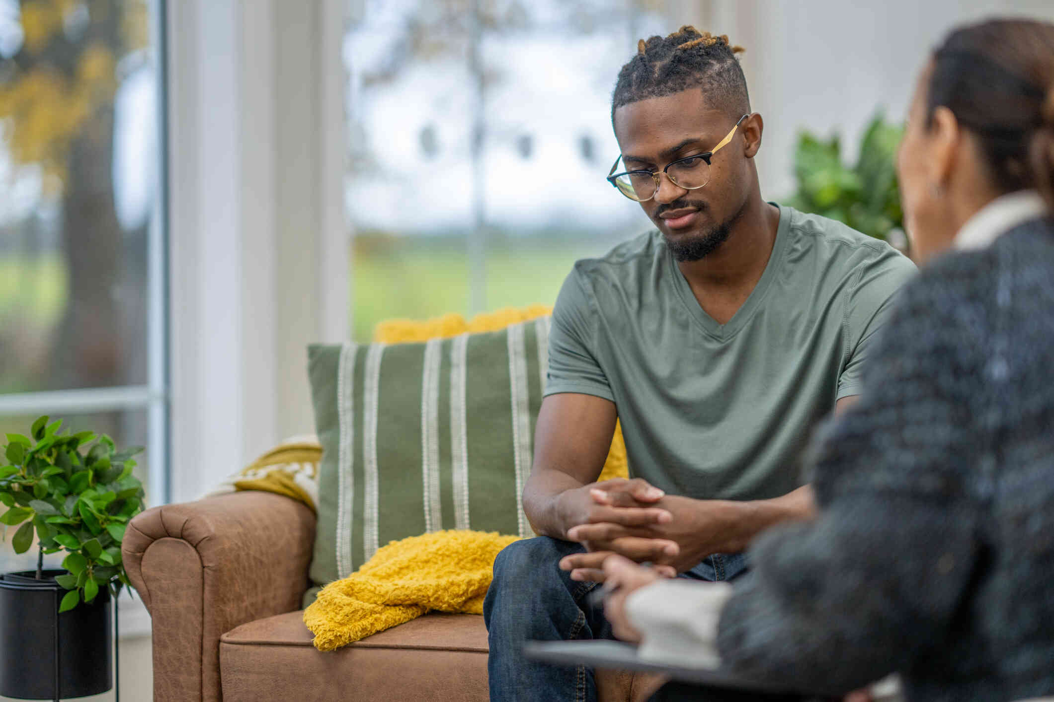 A man in a green shirt and glasses sits hunched over on the couch with his hands pressed together as he listens to his therapist during a therapy session.