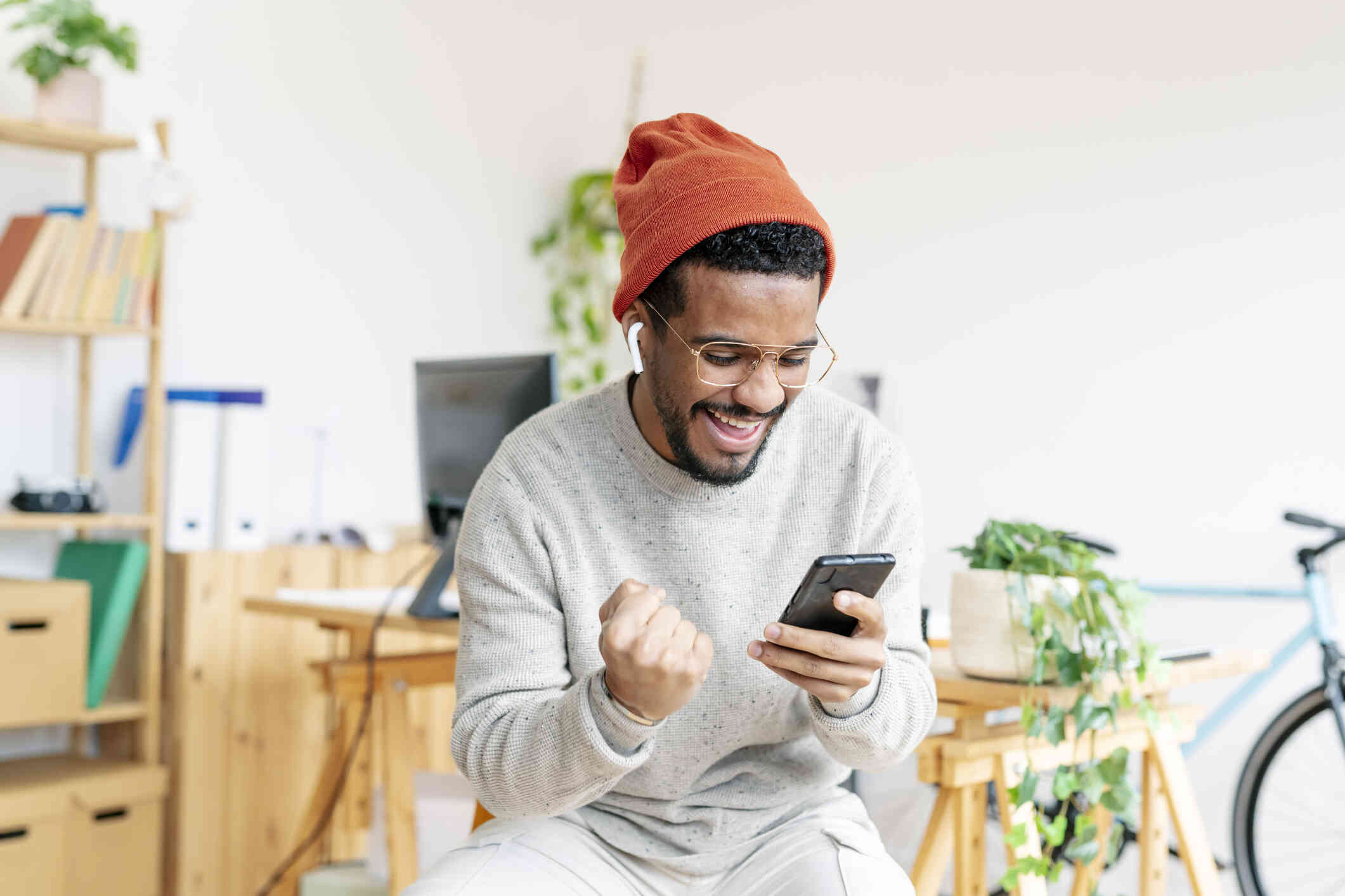 A man in an orange beanie smiles brightly while sitting in his home and looking at the cellphone in his hand.