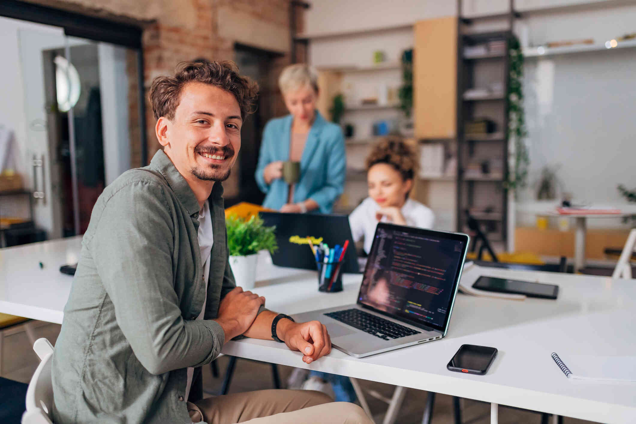 A man smiles as he sits at a table and looks towards the camera. A laptop is open on the table in front of him and two women in the background look at another laptop sitting on the table.