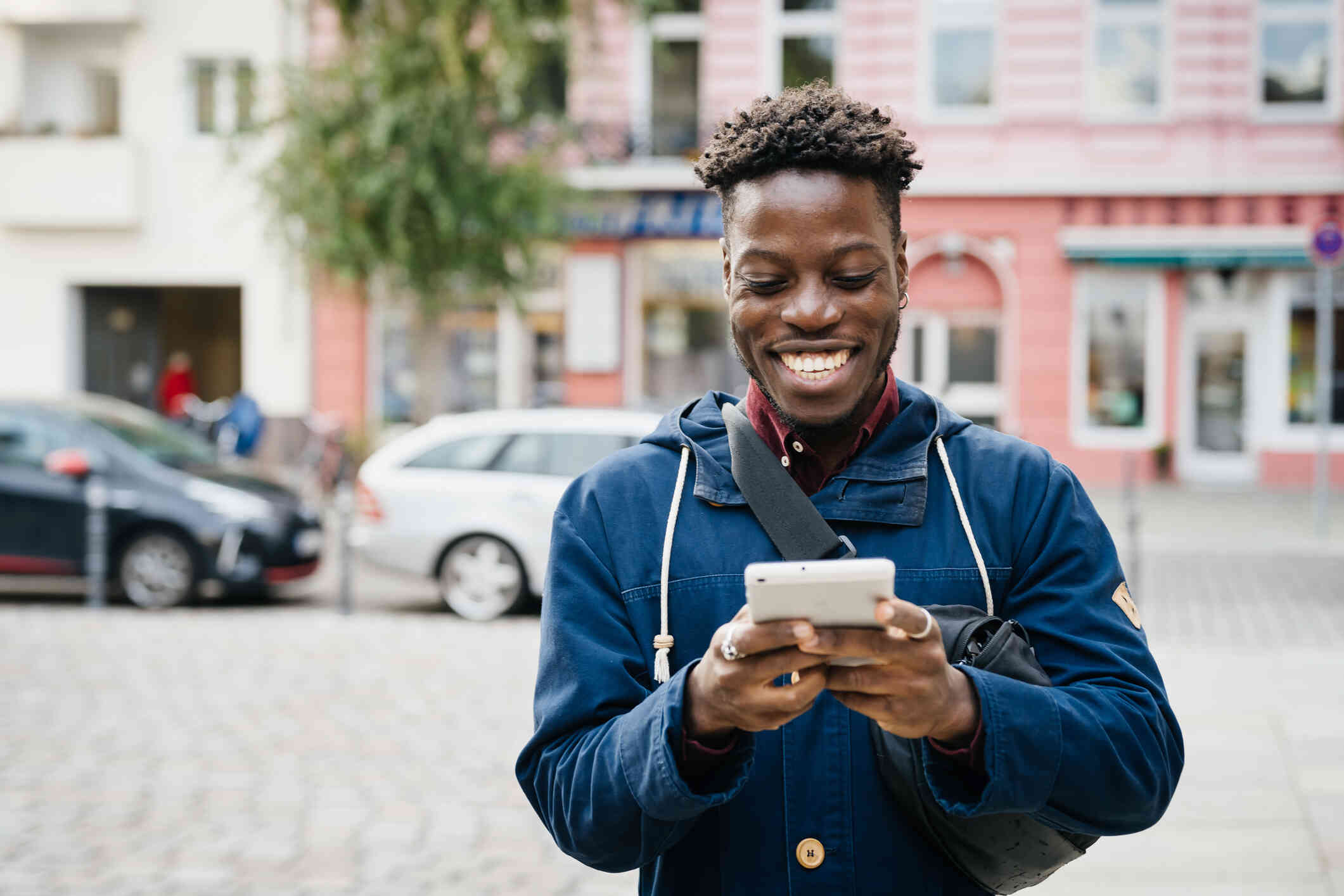 A man in a blue sweater stands outside on a sunny spring days and smiles brightly while looking down at the phone in his hand.