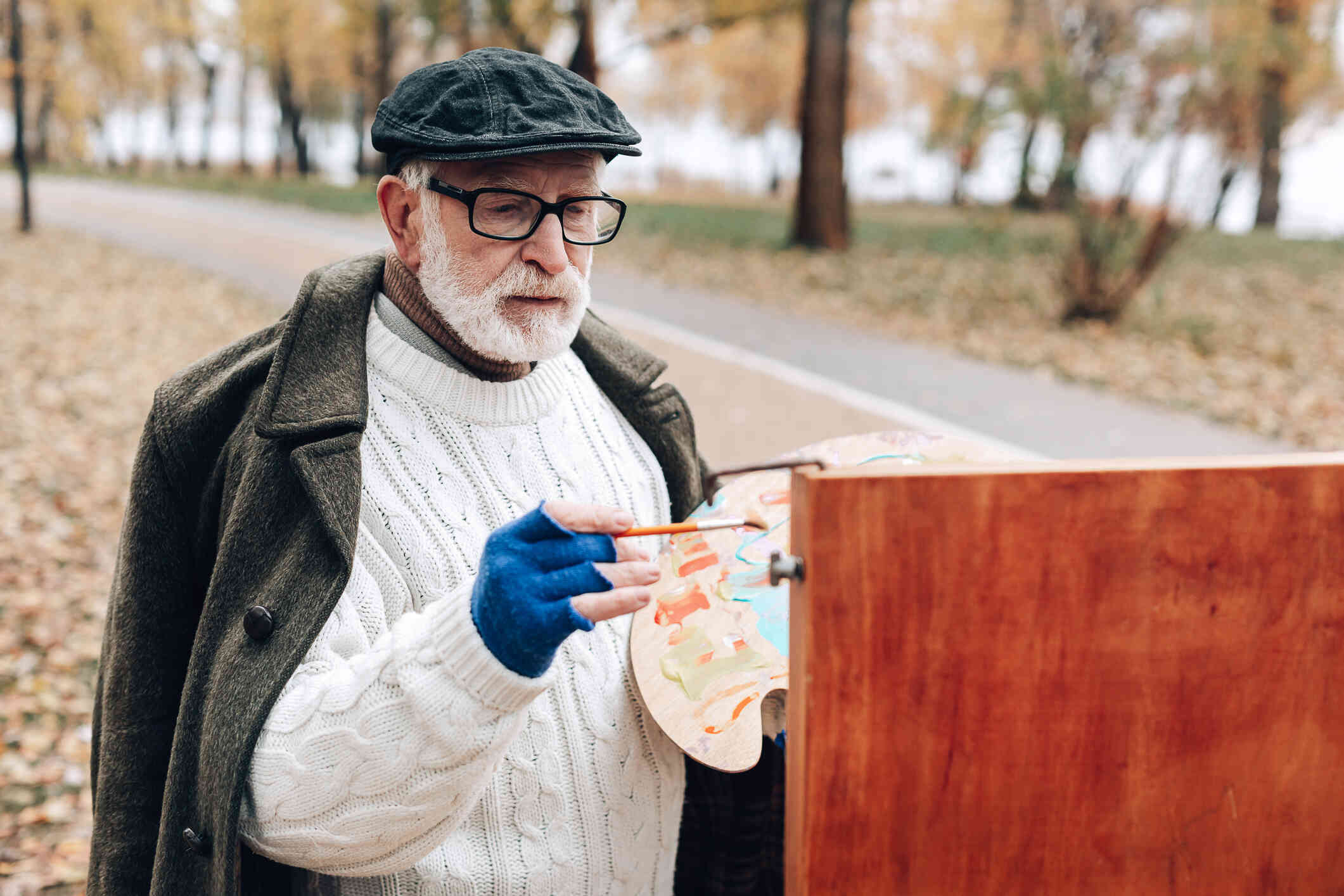 An older man in a sweater and hat stands outside with his painbrush and paints on a canvas on an easle.