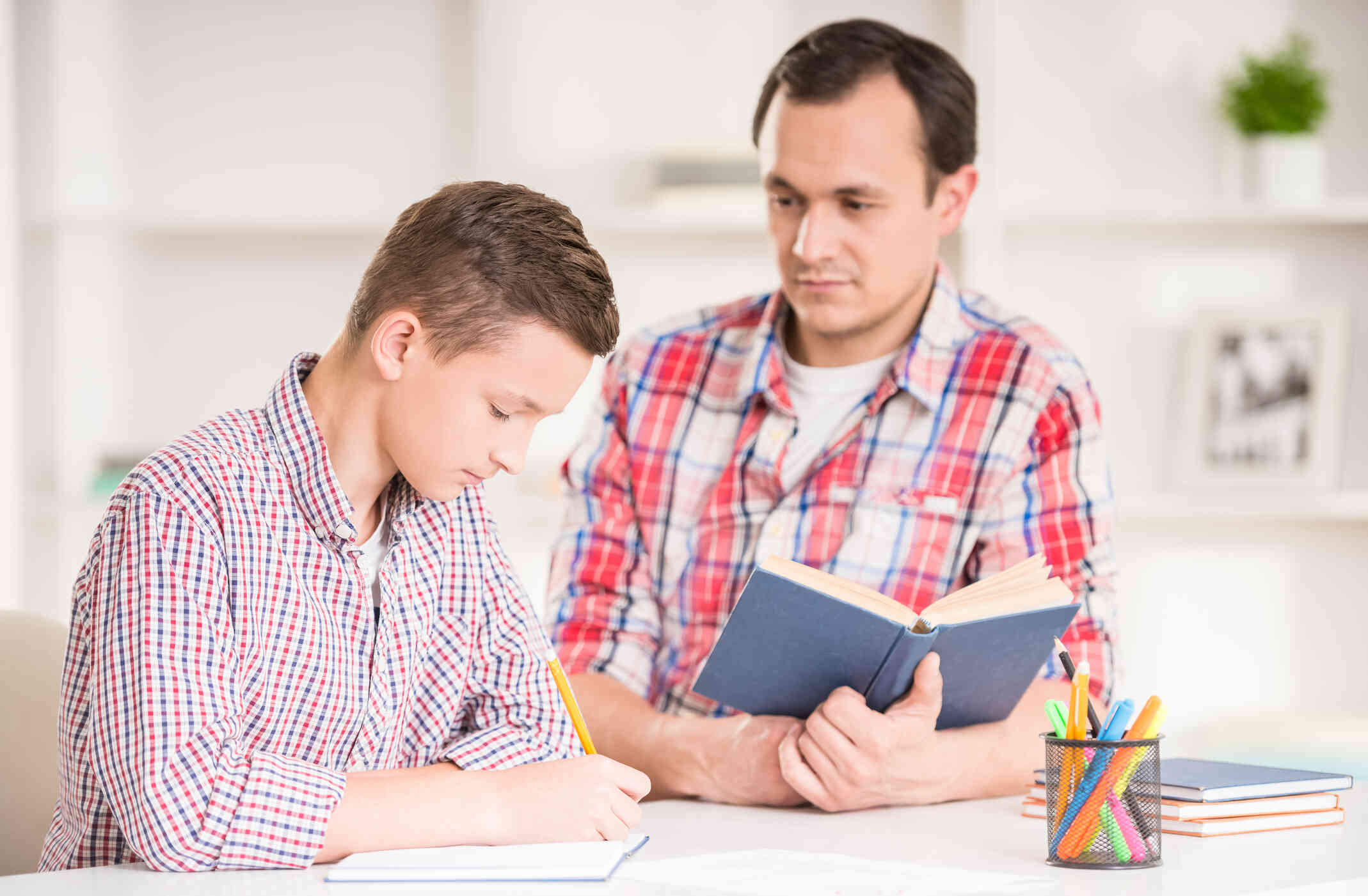 A man sits at a table with an open book as he looks at his son who sits next to him while doing homework.