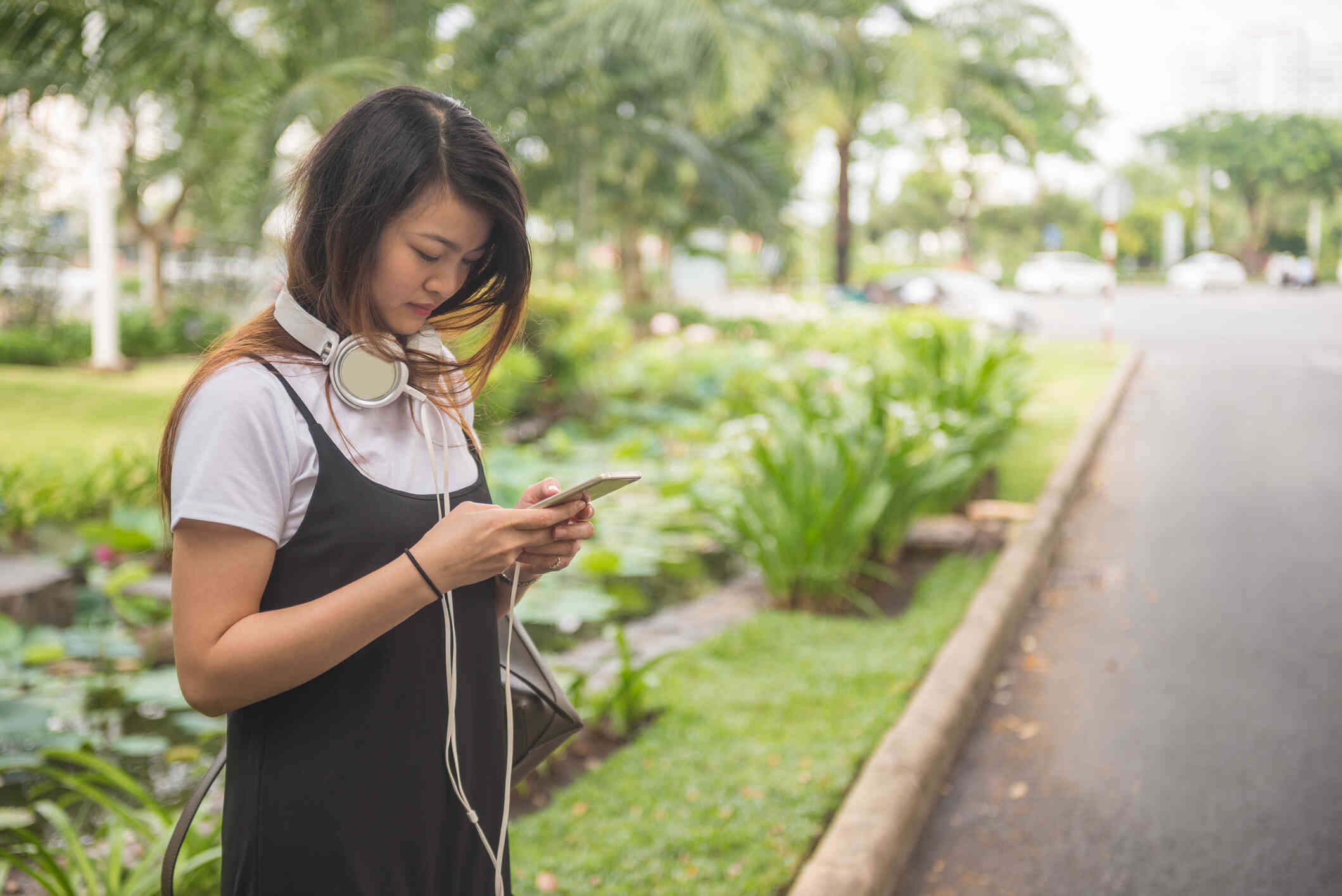 A teen girl in a black dress and white headphones stands outside and looks at the cellphone in her hands.