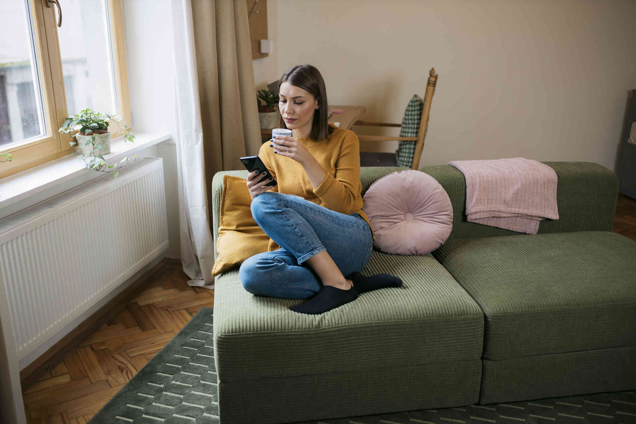 A woman in a yellow shirt sits curled up on a green couch with a cup of coffee while looking at the cellphone in her hand.