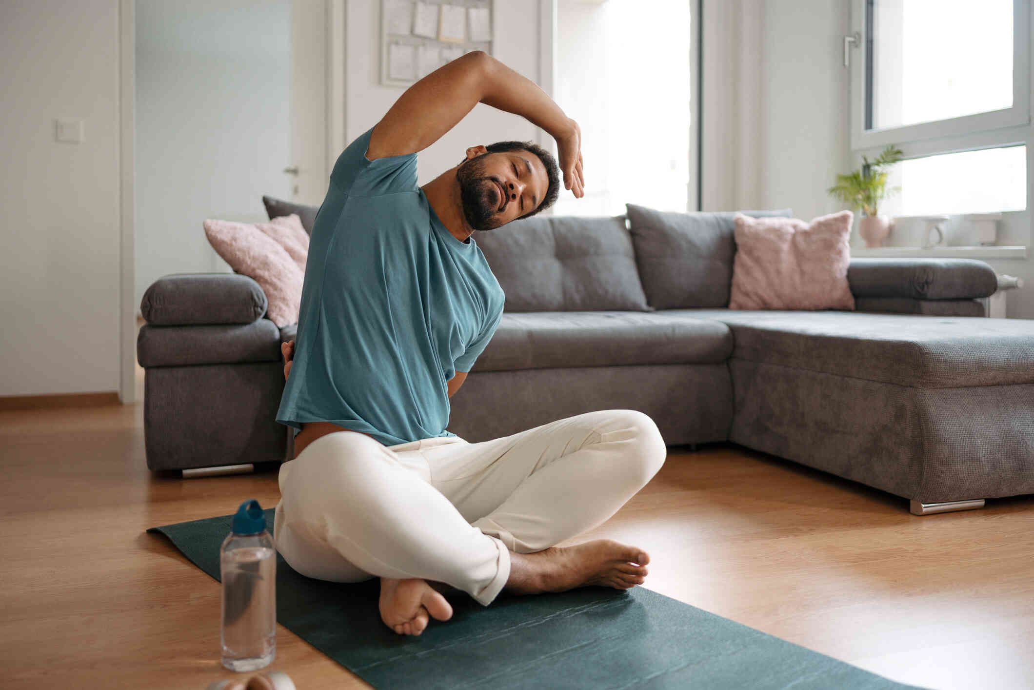 A man in a blue shirt sits on a yoga mat while doing a stretching pose.