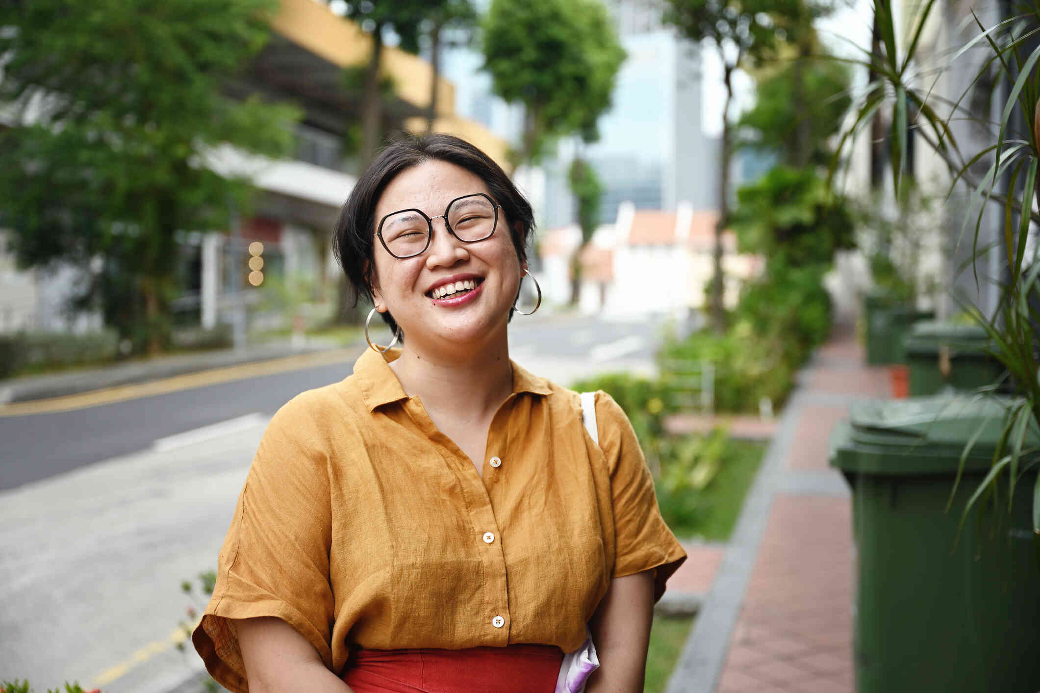 A woman wearing glasses and an orange blouse smiles wide as she stands outside on a sidewalk.