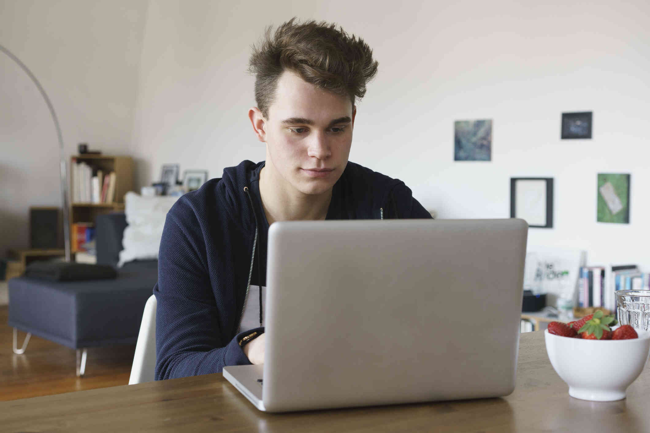 A young man sits at a table in his house looking at his computer with a neutral expression