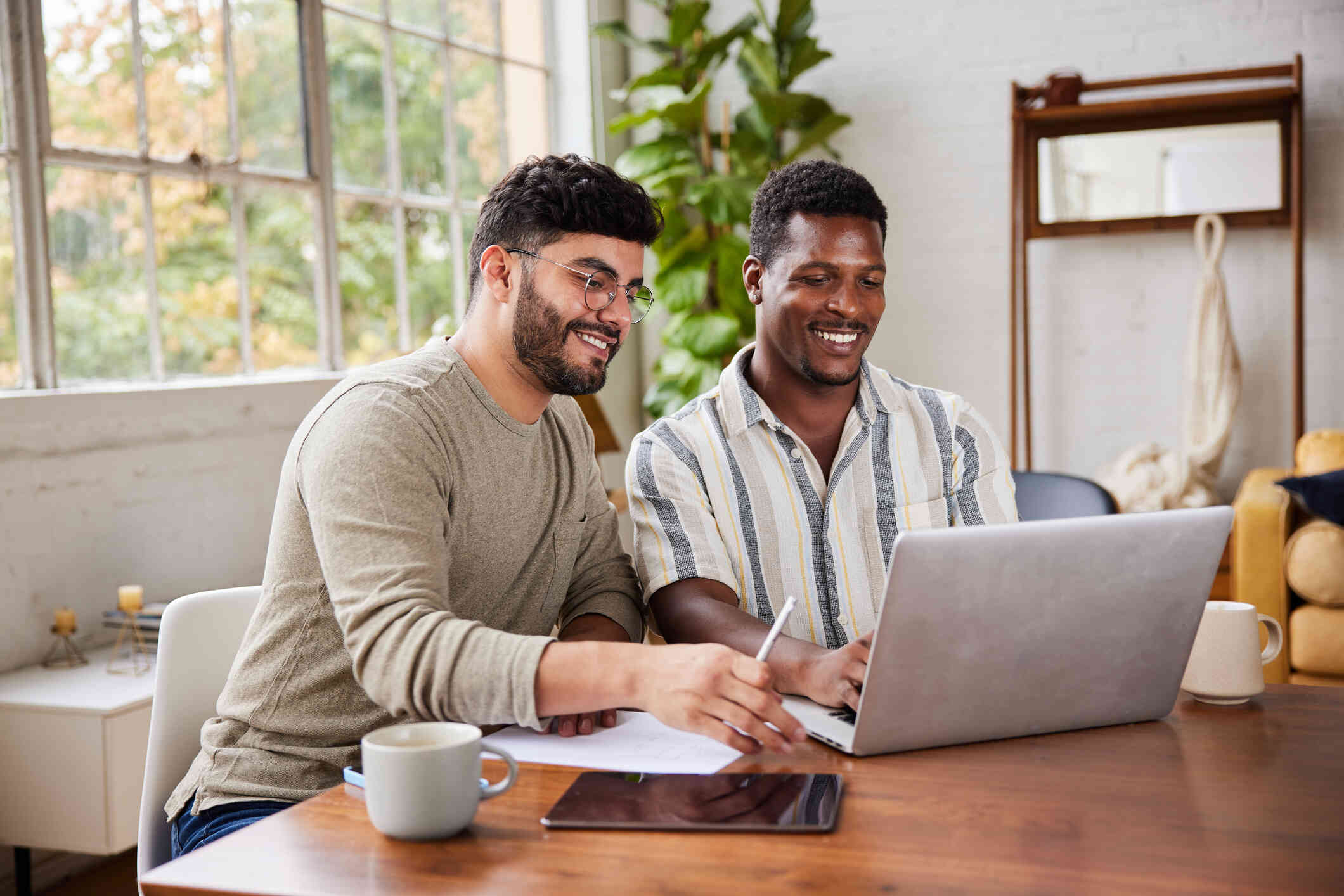 Two men smile as they sit at a kitchen table and look towards a laptop screen sitting in front of them.