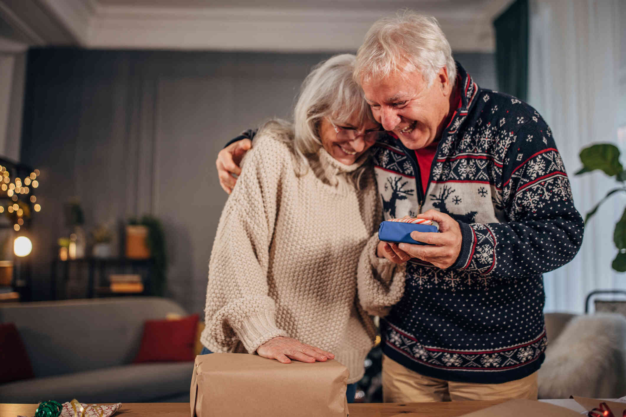 A mature man and woman smiles as they stand and hug each other while the man holds a wrapped gift with a candy cane on top of it.