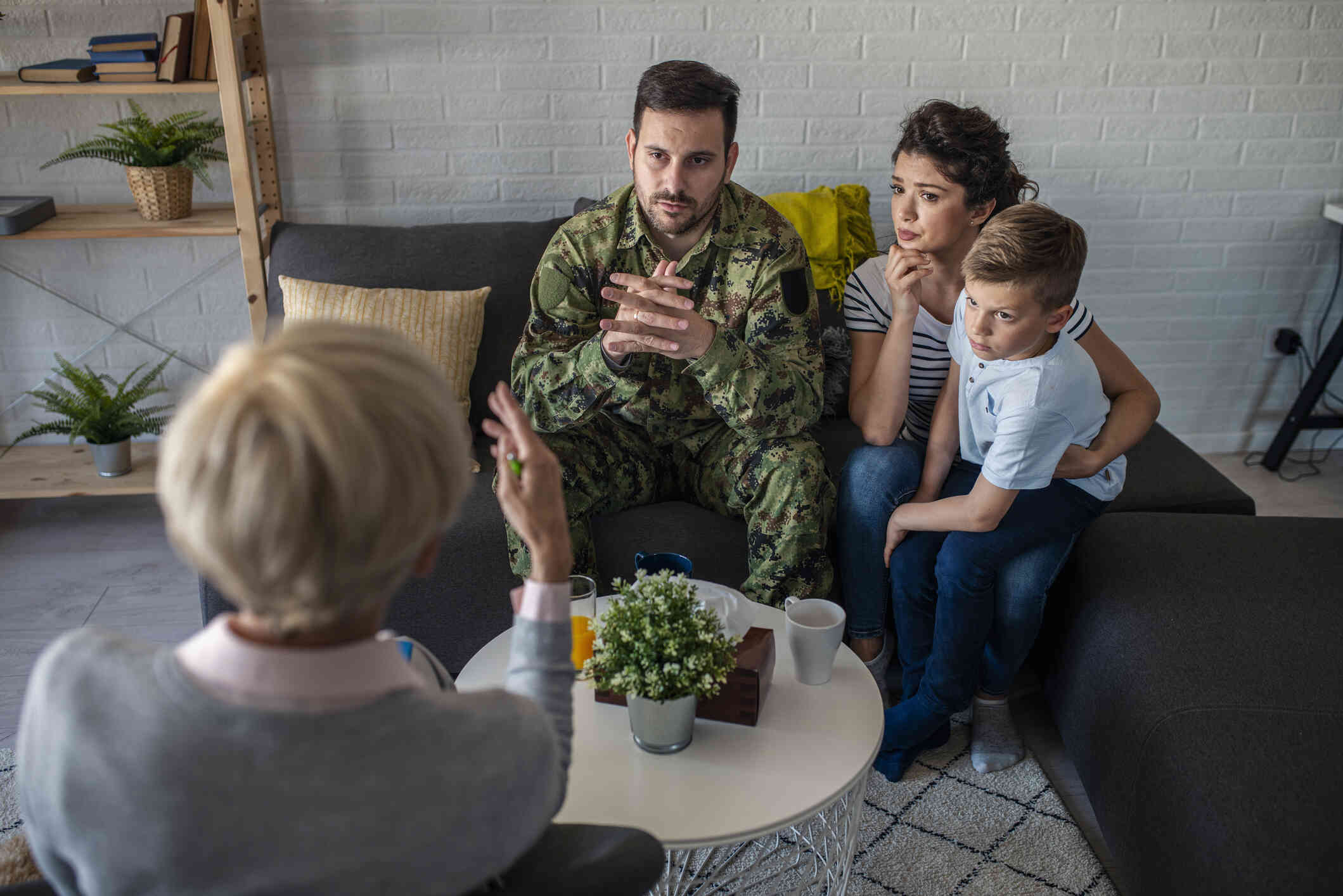 A young buy sits on his mothers lap as they sit on the couch next to his father in a military uniform and talk to the therapist sitting across from them.