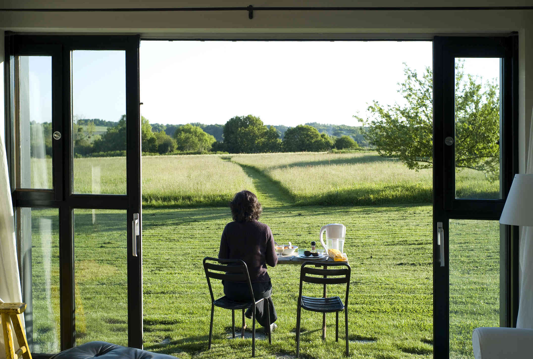 A woman with her back to the camera sits outside at a table iwth food and gazes out of the lush rolling hills infront of her.