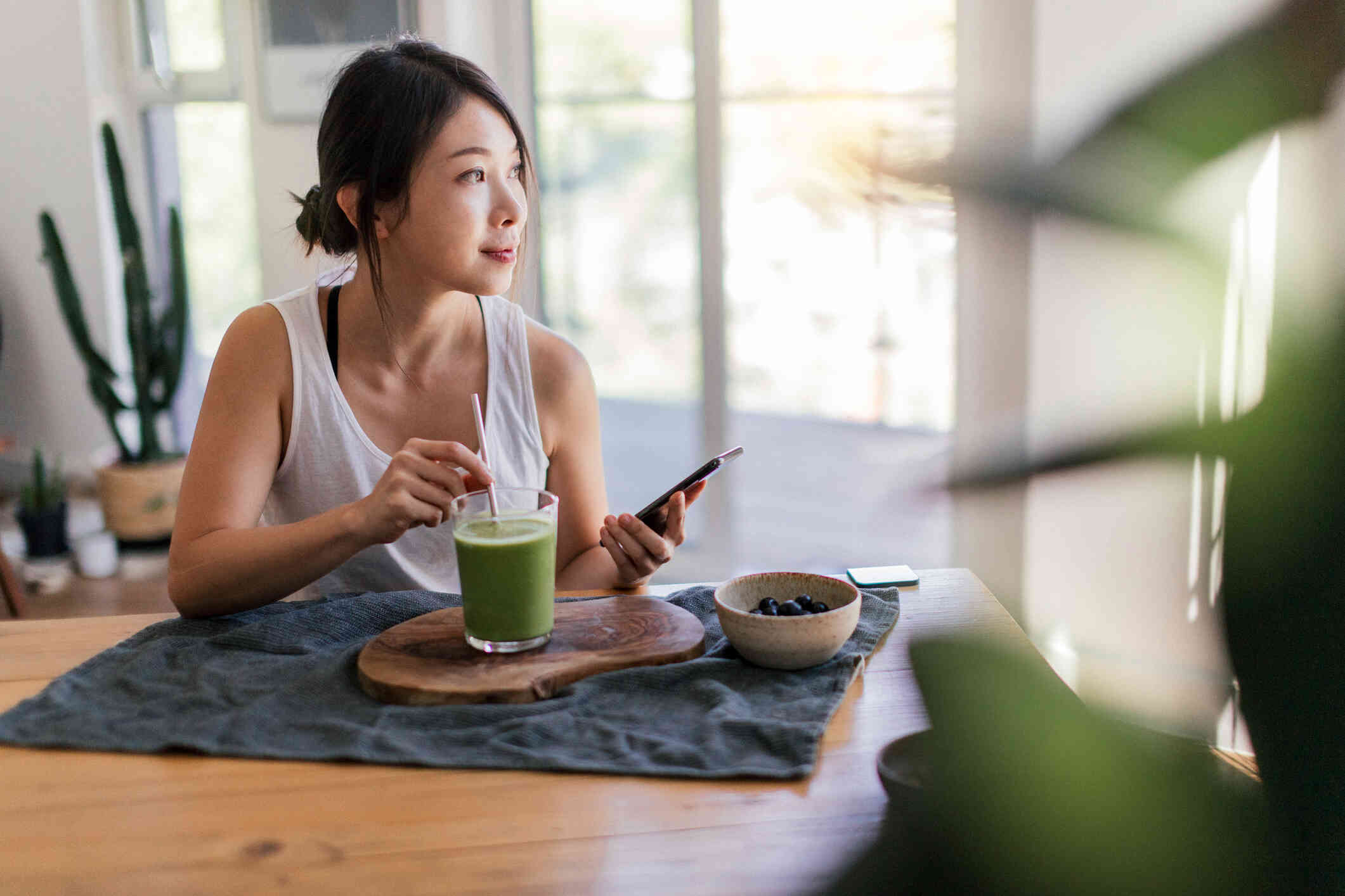 A woman in a white shirt sits at the kitchen table with a green smoothie as she holds a phone in her hand and gazes off with a slight smile.
