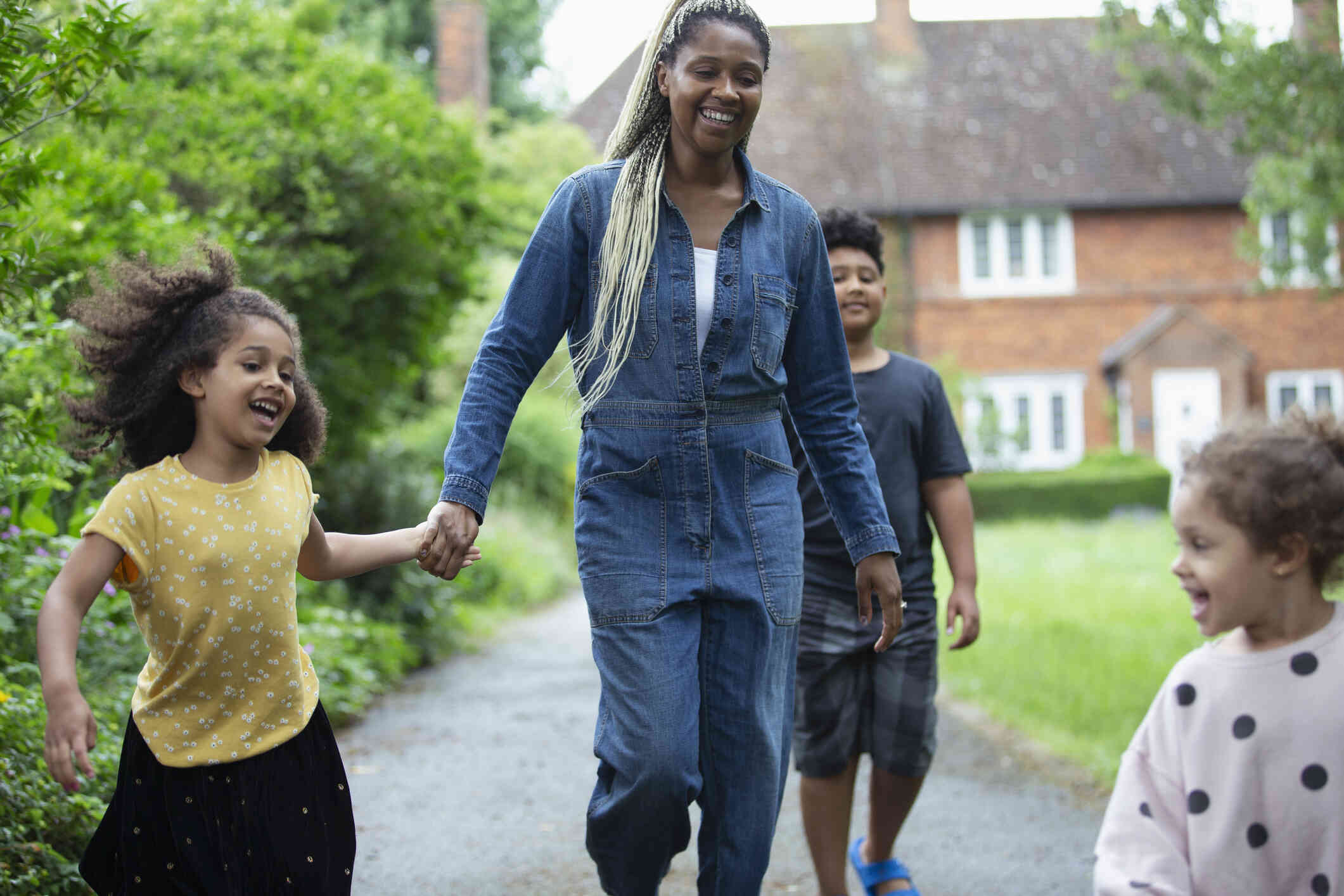 A mother in blue jean coveralls walks walks outside with her son and daughter on a sunny day while smiling.