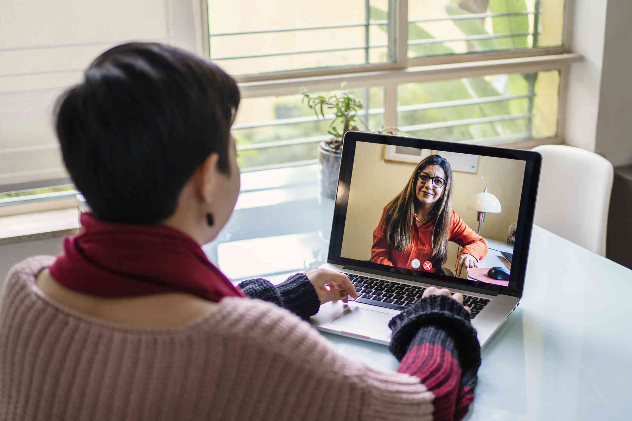 A close up of a woman sitting at a table near a window as she talks to the female therapist on the laptop screen open infront of her during a virtual therapy session.