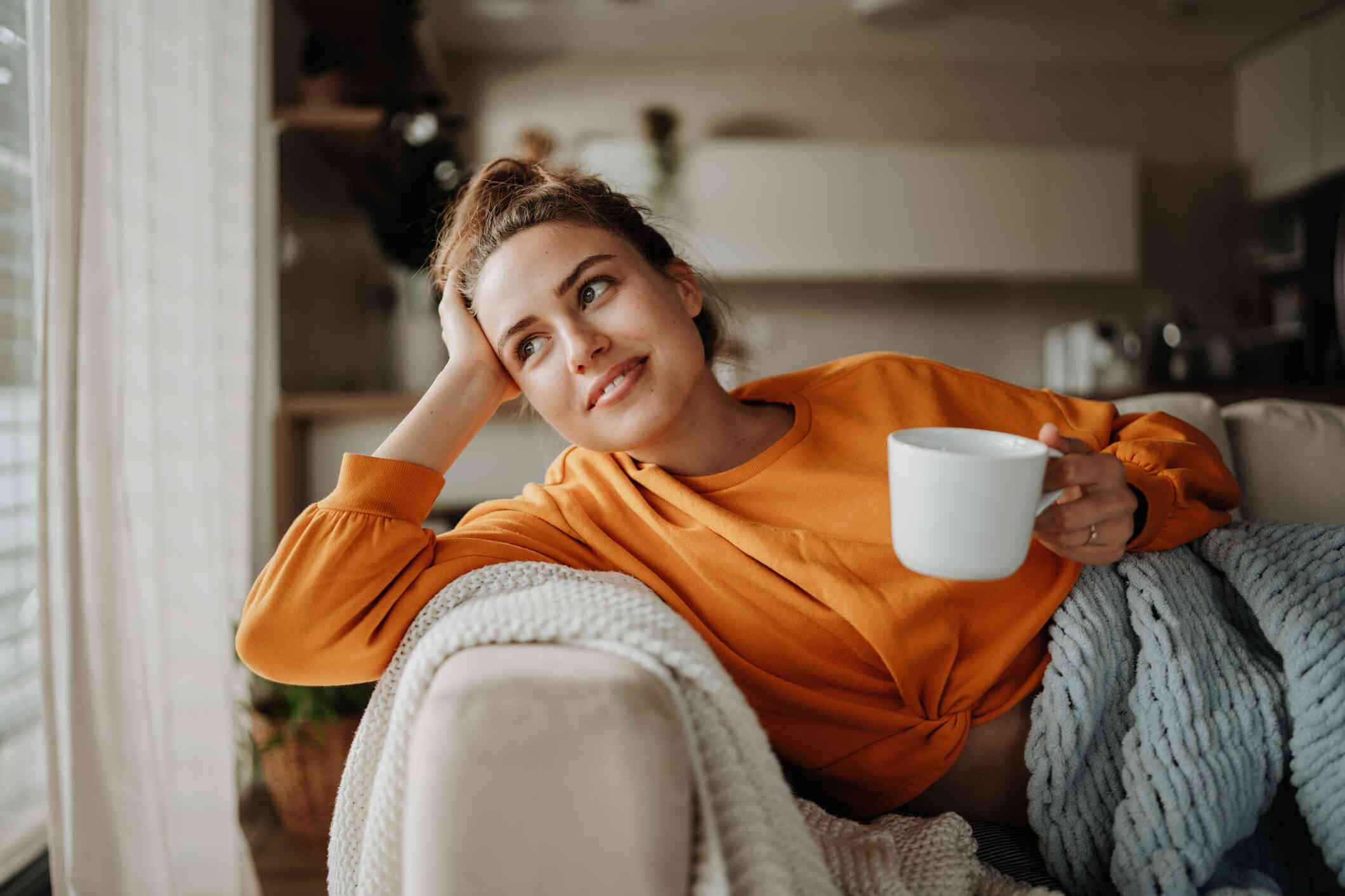 A woman in an orange shirt reclines on a chair with a cup of coffe and smiles softly while gazing out of a window.