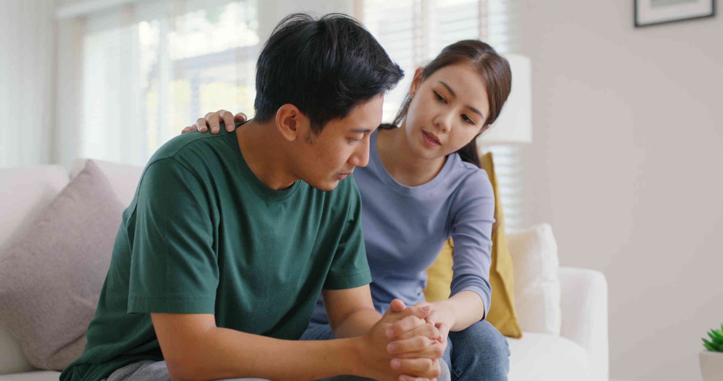 A man sits sadly hunched over on the couch as his female partner sits next to him an tries to comfort him.