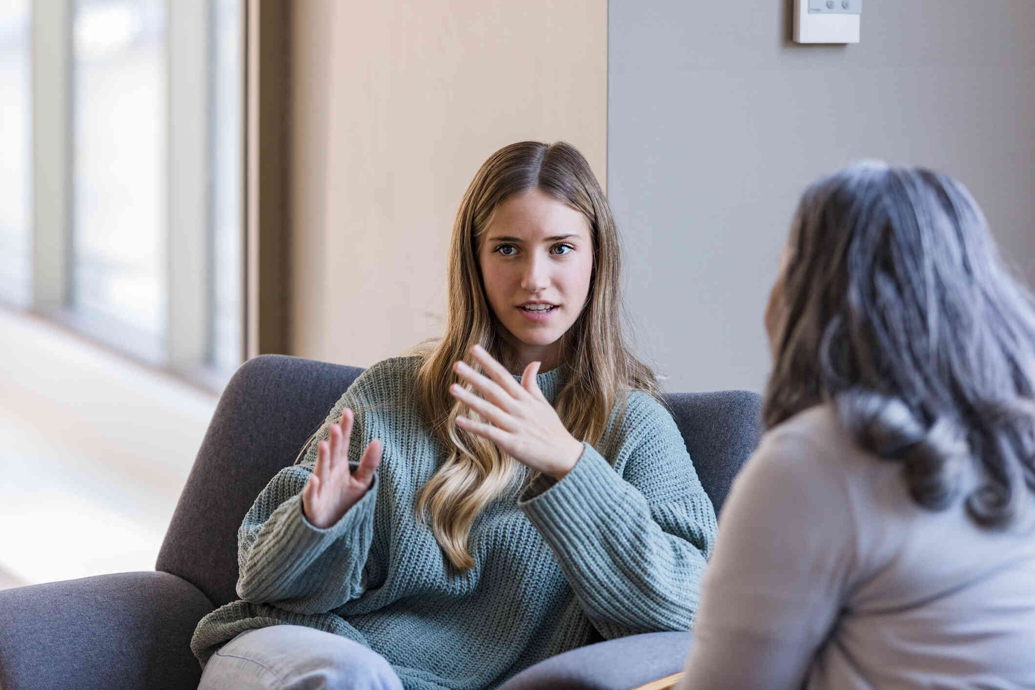 A young woman in a green sweater sits on a couch while speaking with hand gestures with a counselor in a gray top