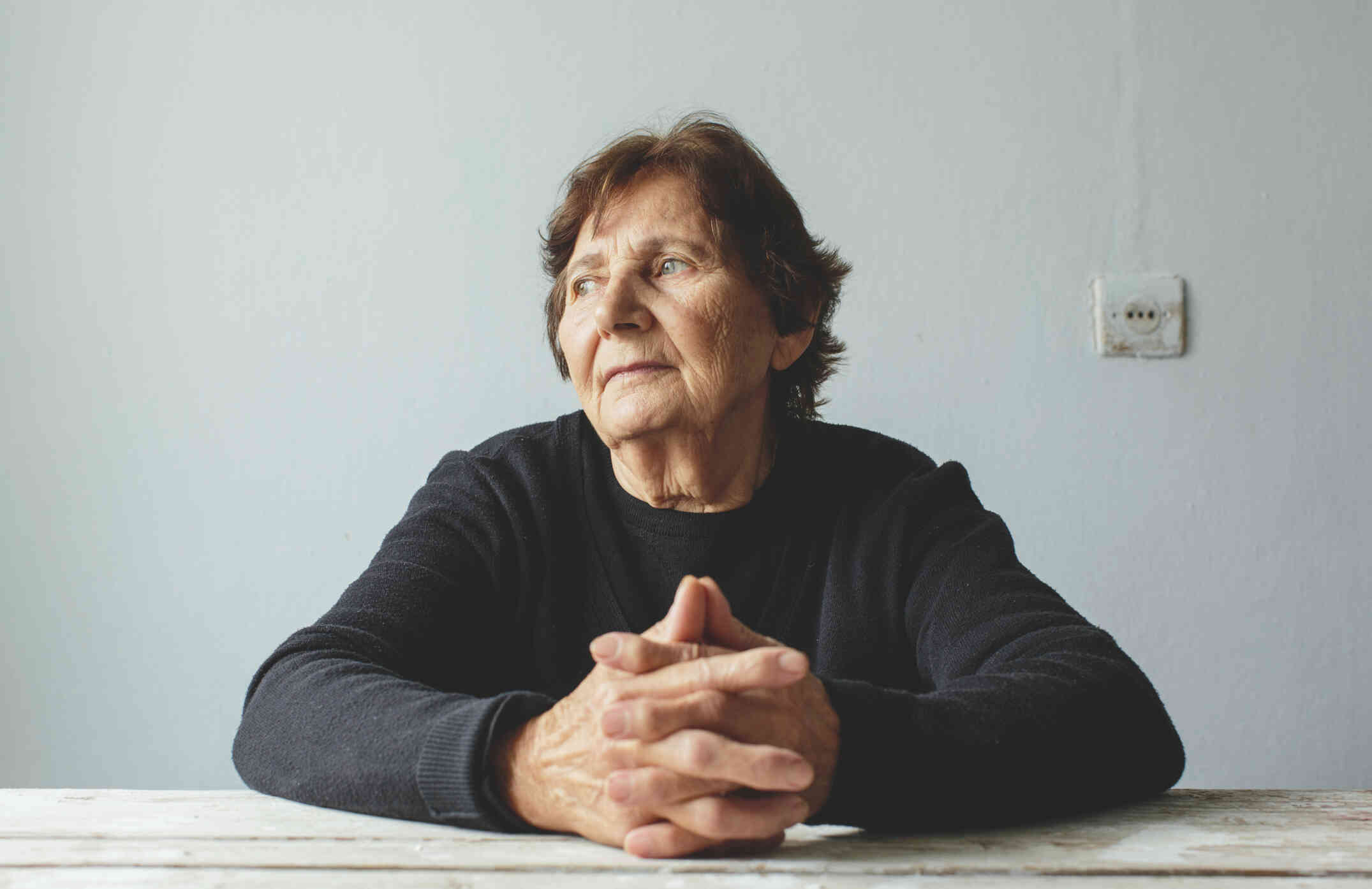 A mature woman in a black shirt sits at a table with her hands pressed together as she gazes off sadly.