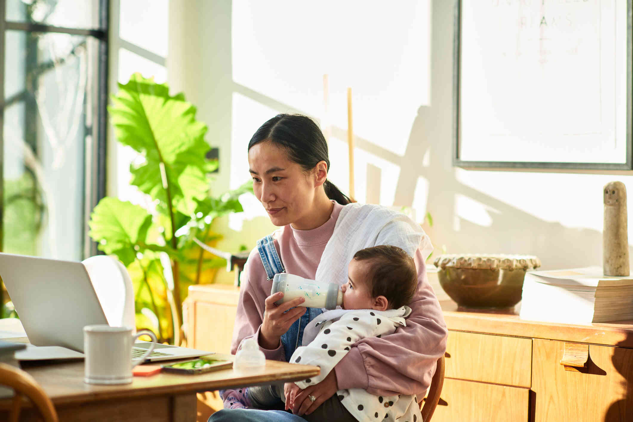 A mother with a burp cloth on her shoulder feeds her baby from a bottle while sitting at a table and looking at the laptop open infront of her with a serious expression.