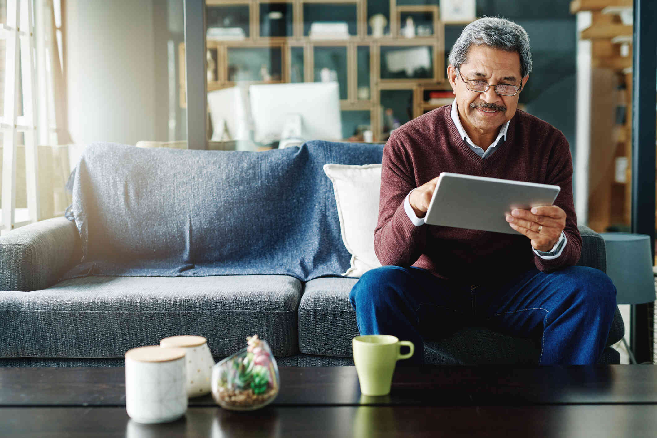 An older man with glasees sits on his couch and looks at the tablet in his hands with a smile.