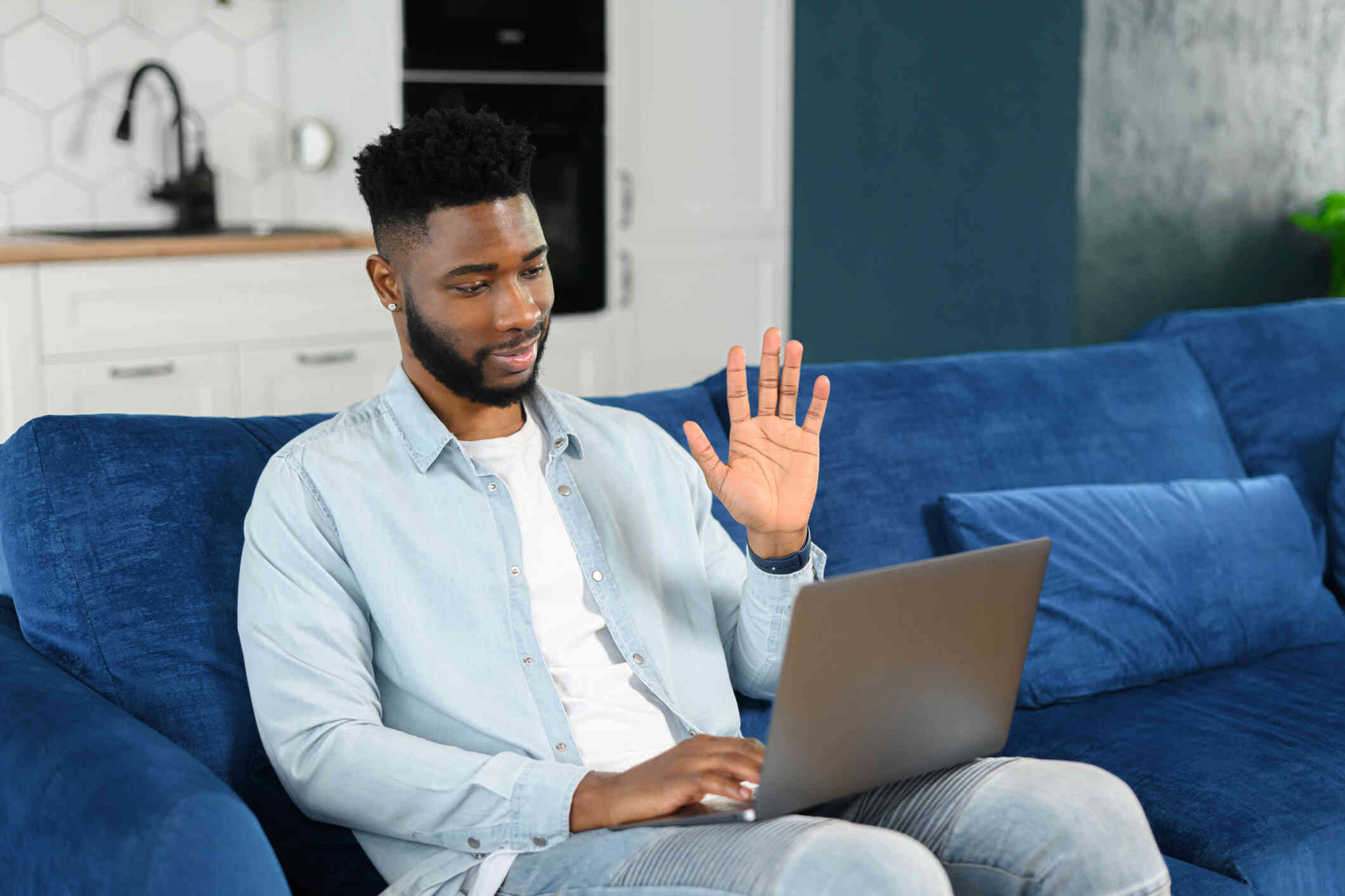 A teenage boy seated on a blue couch waves and smiles at his laptop screen.