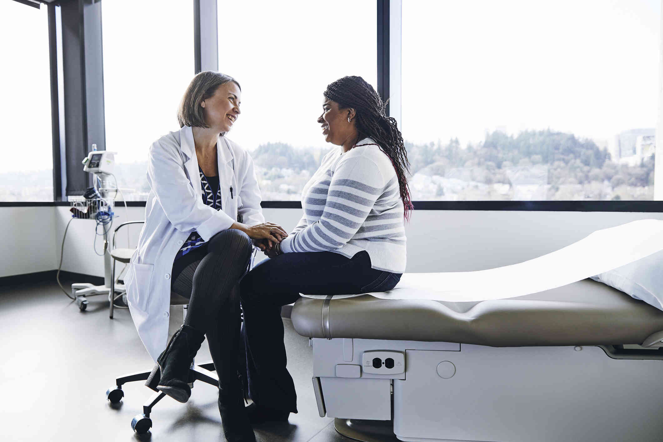 A woman in a striped sweater smiles as she sits on the edge of a hospital bed and a female doctor in a white coat sits next to her and speaks.