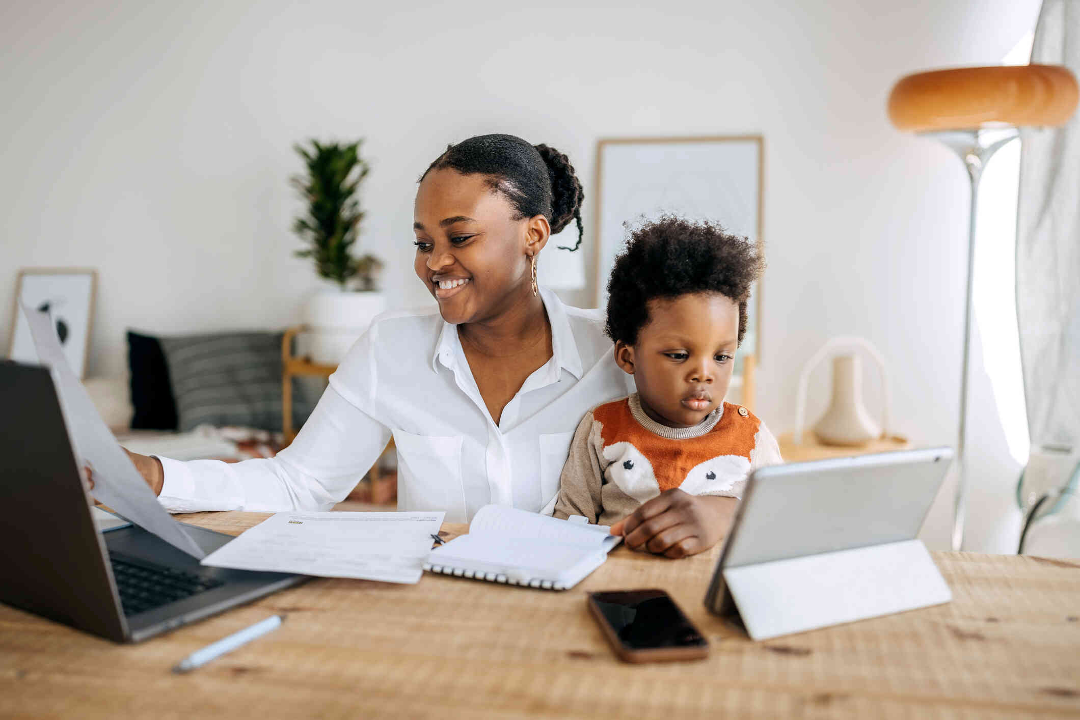 A mother in a white blouse sits at the table and smiles at some papers in her hand while her young son sits in her lap and looks at the tablet on the table.