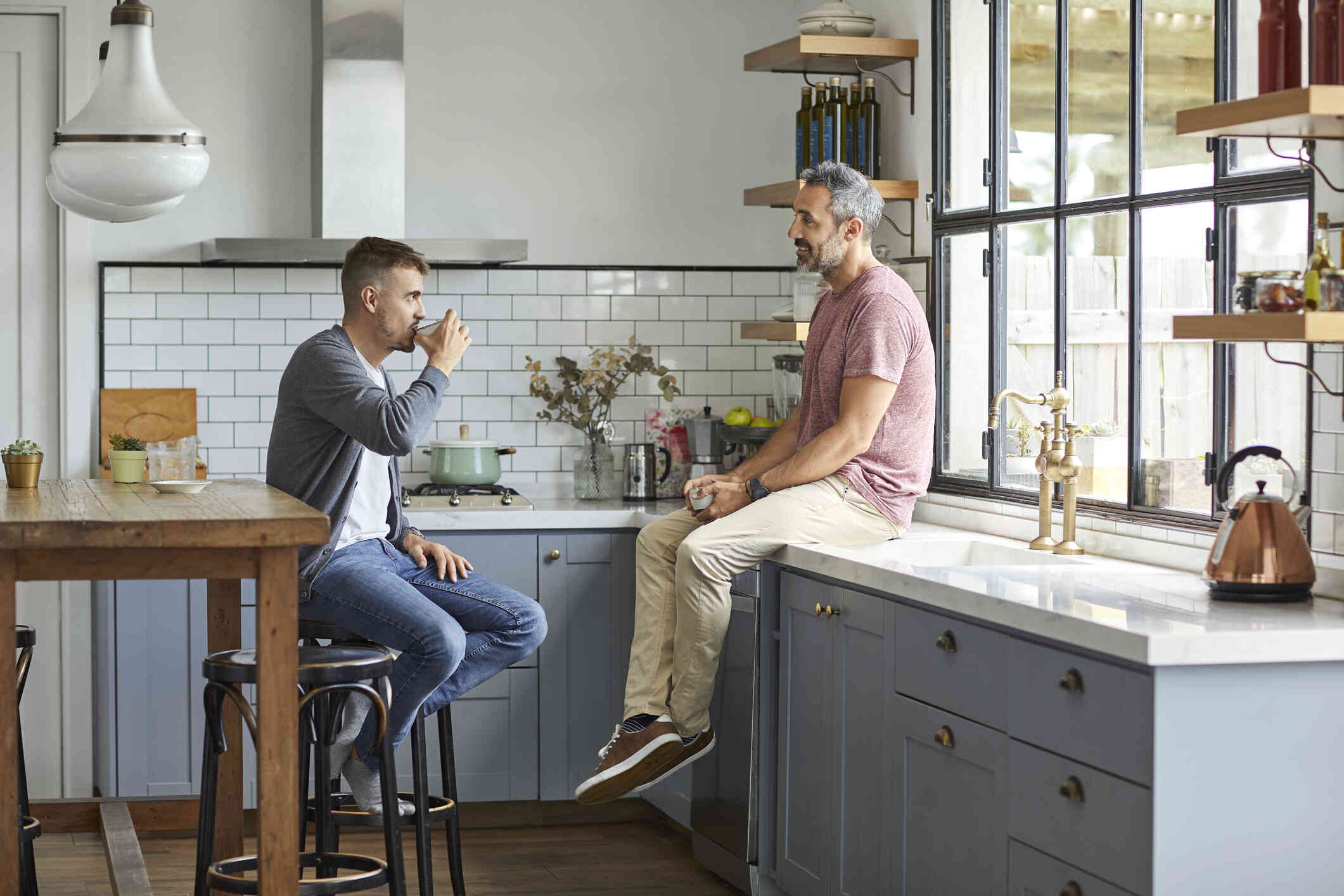 A man sits on the kitchen counter with a cup of coffee while his male partner sits across from him on a stool as they talk.