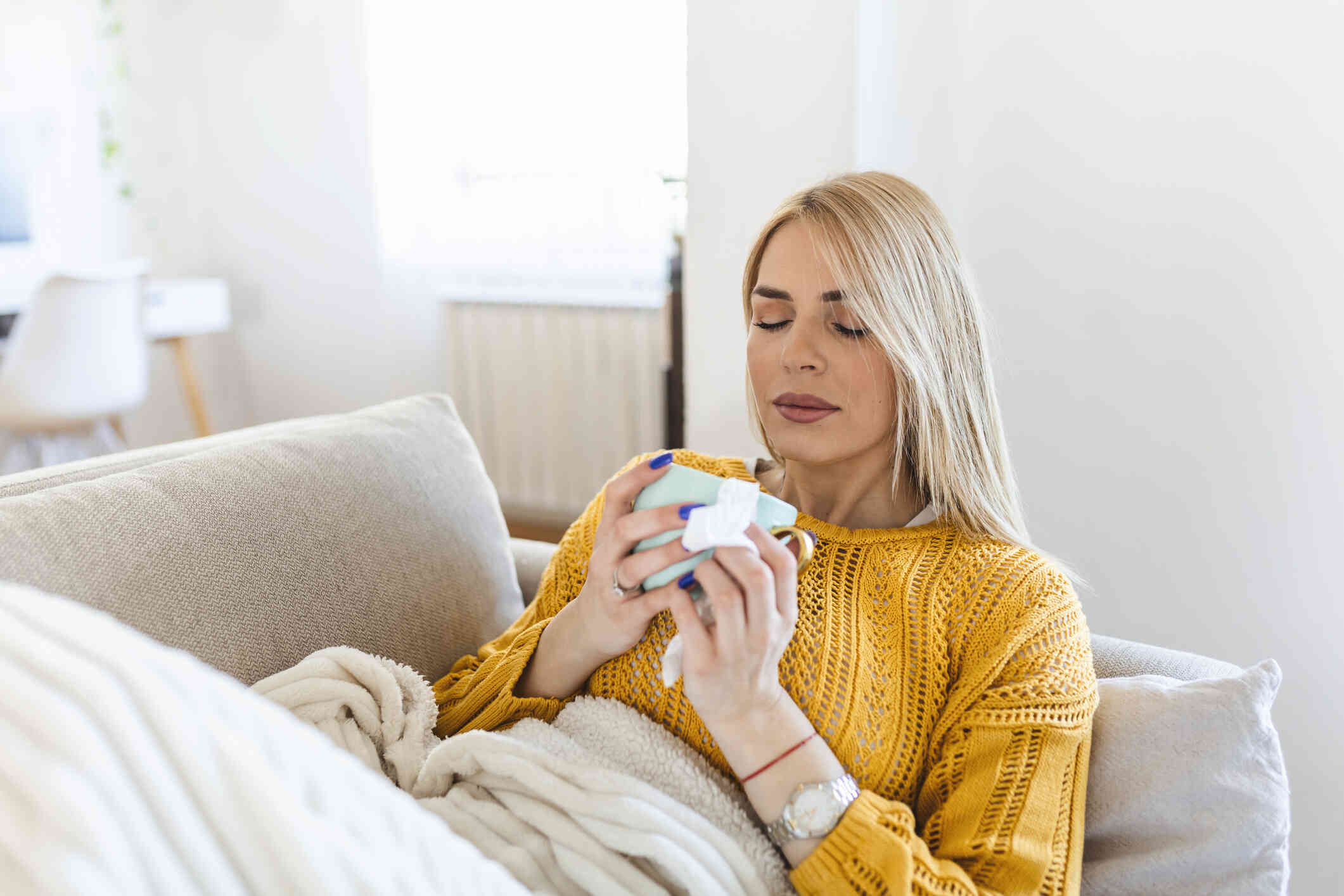 A woman in an orange sweater reclines on the couch under a blanket while holding a coffee mug and closing her eyes.