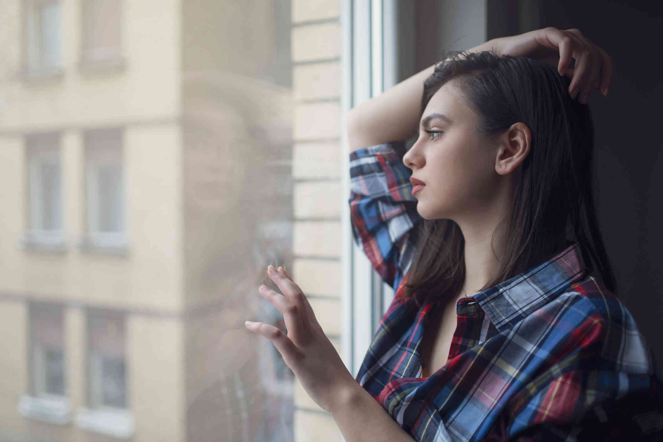 A woman in a plaid shirt gazes out if window while resting her fings on the glass with a sad expression.