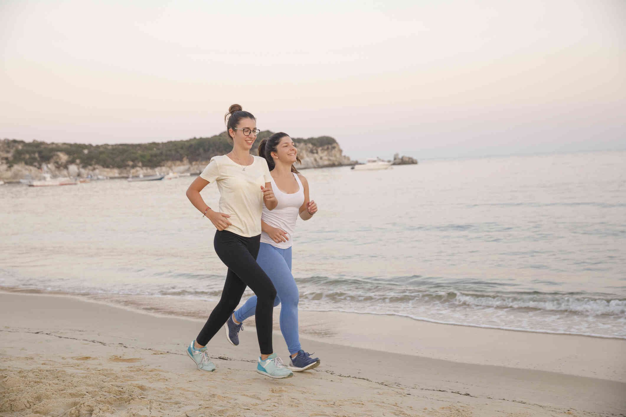 Two female friends in workout clothes jog together down a beach on a sunny day.