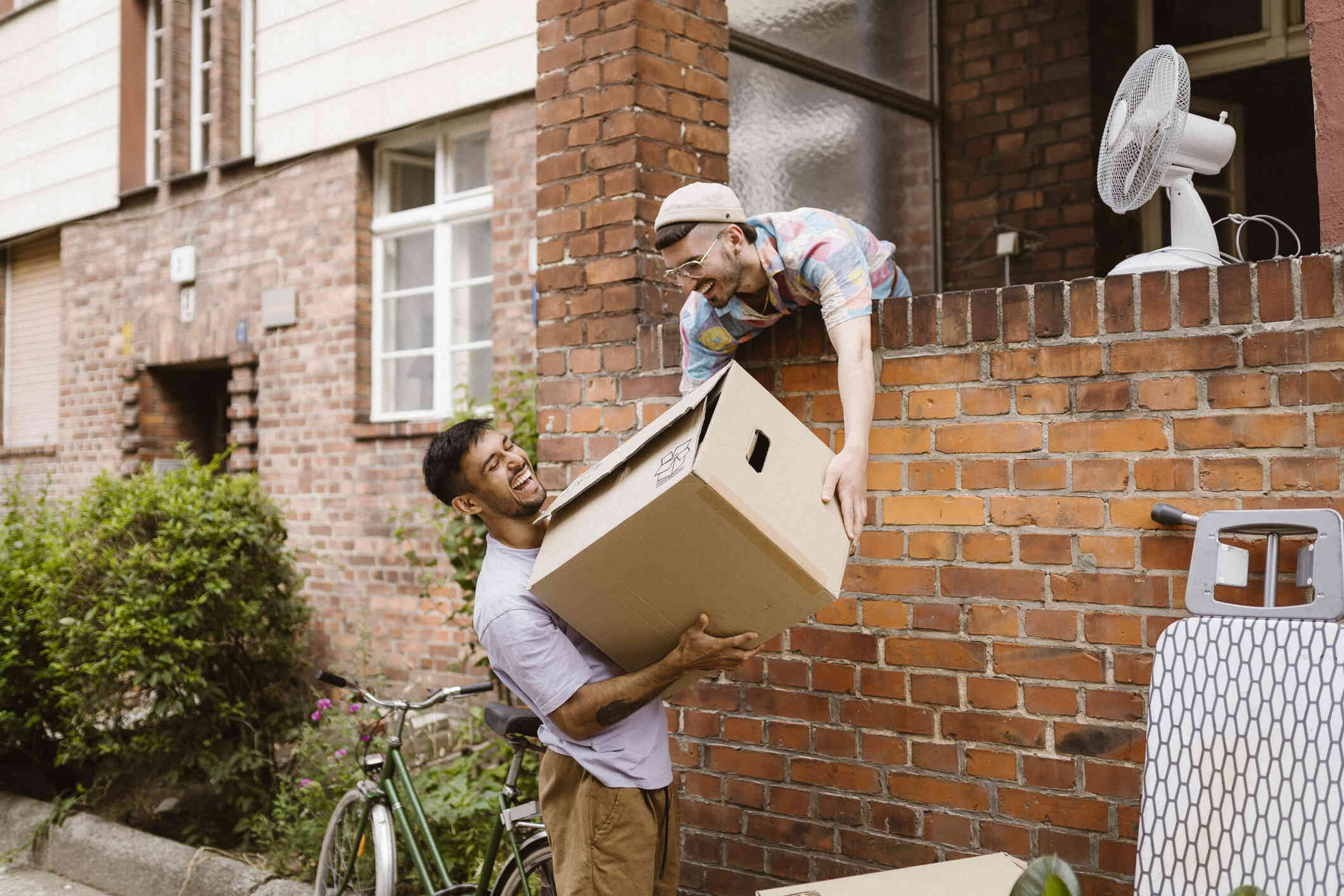 A young man in a hat helps hand over a cardboard box to another man wearing a grey shirt as they smile