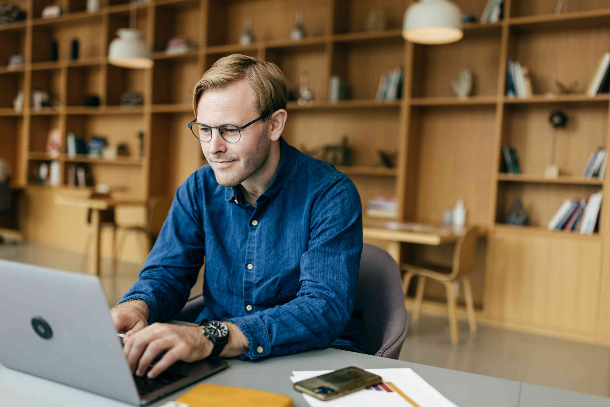 A man in a blue botton down shirt sits at a table and types on the laptop open on the table infront of him.