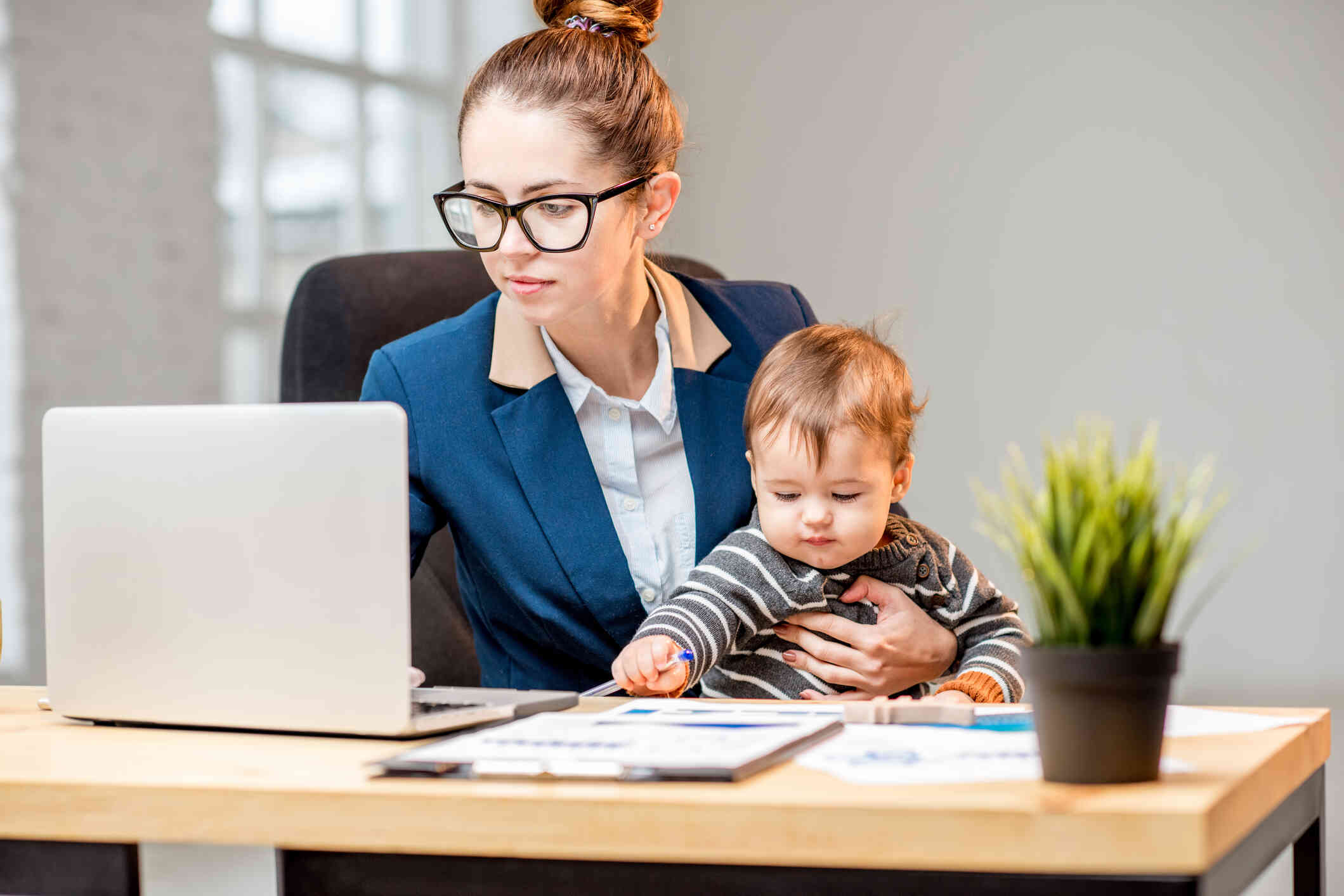 A mother wearing a blue blazer and glasses sits at a table while working on her laptop as she holds her toddler son in her lap.