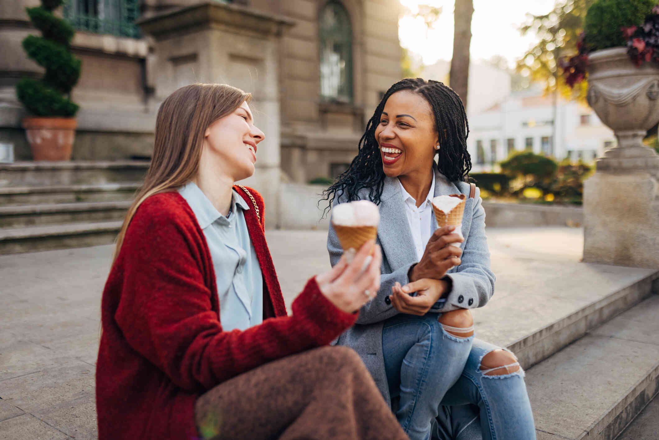 Two adult female friends sit next to each other outside on a sunny day while eating ice cream and laughing.