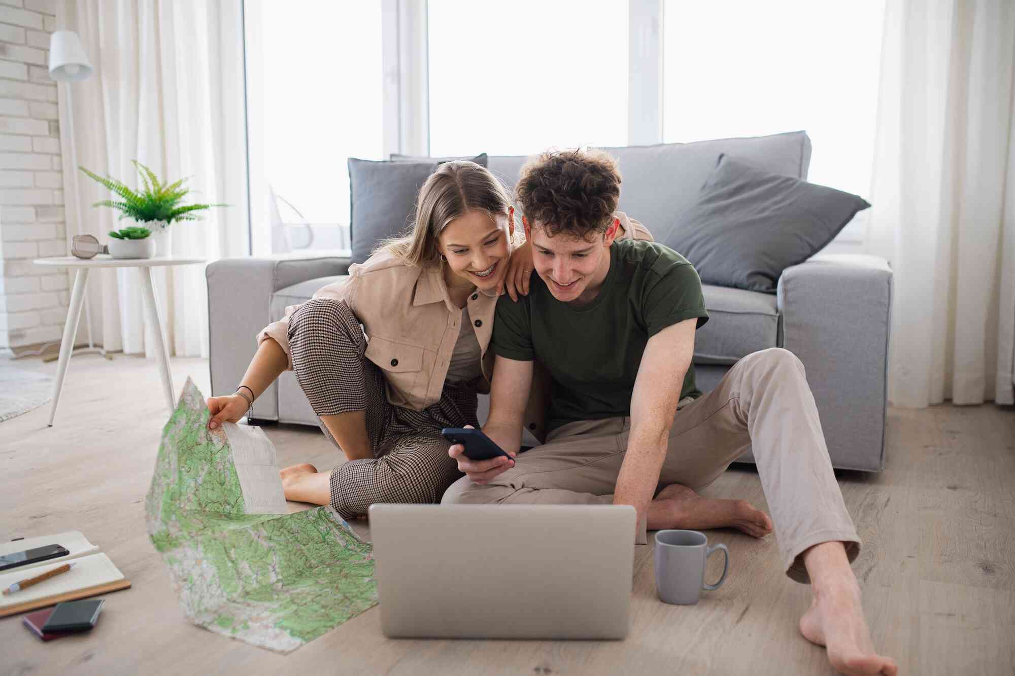 Two teenagers sit on the floor, both looking at the laptop screen while the girl holds a map.