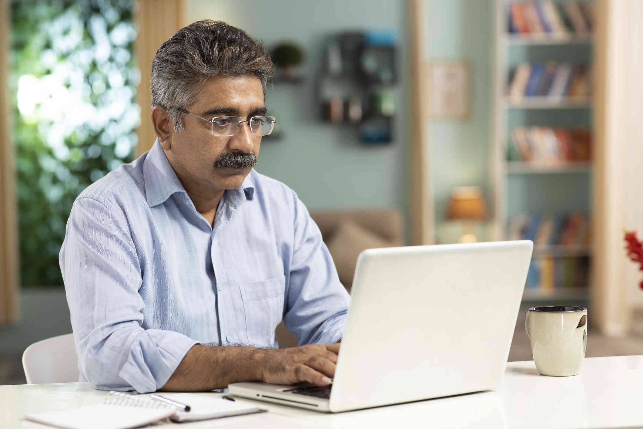 A middle aged man with glasses sits at a wooden table and types on the laptop open infront of him.