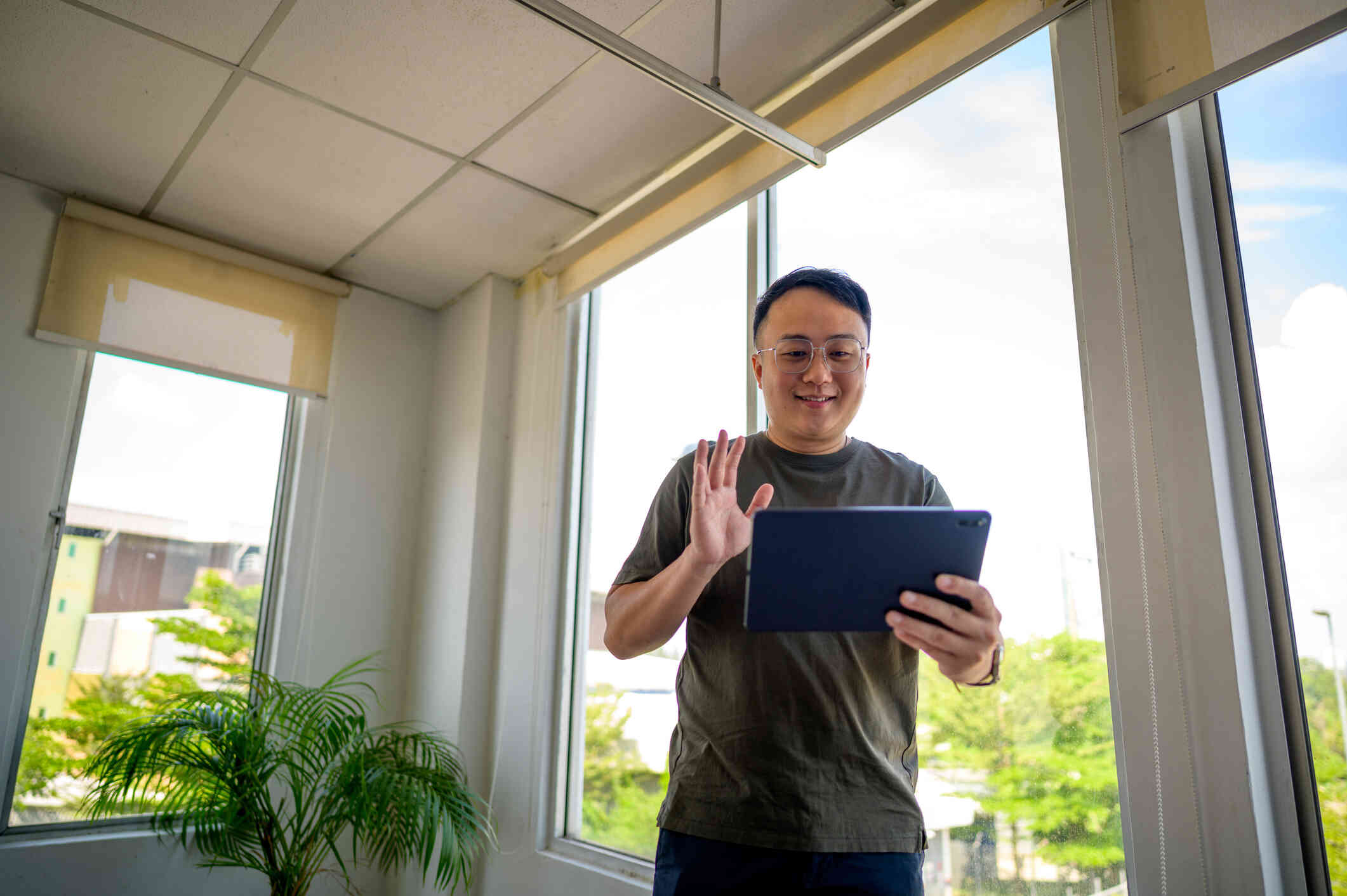 A man with glasses smiles as he stands by a large window and holds a tablet in his hand. He waves towards the screen with his other hand.