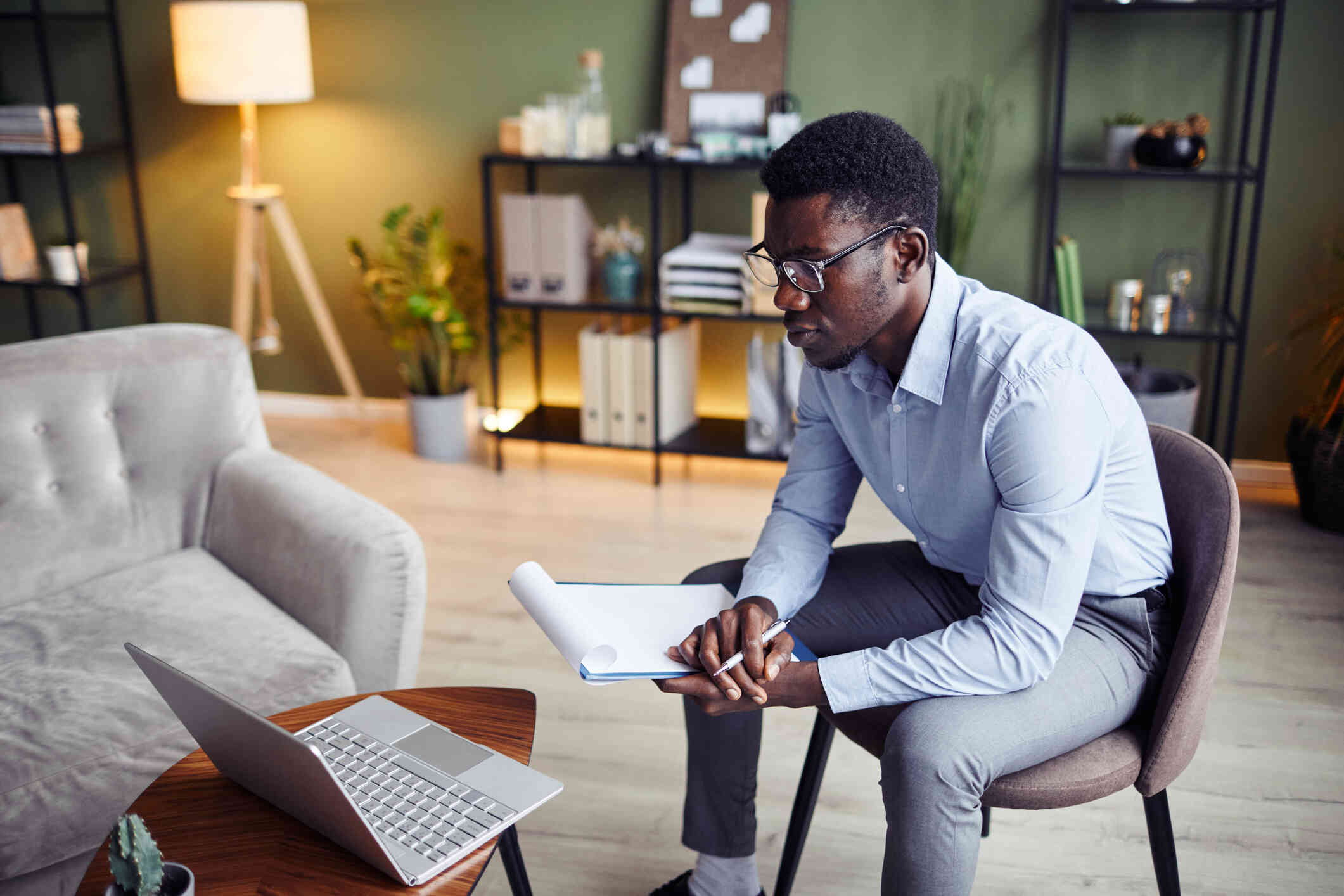 A man in a button down shirt leans forward in his chair while holding a notepad and pen as he looks at the laptop open on the coffee table infront of him.